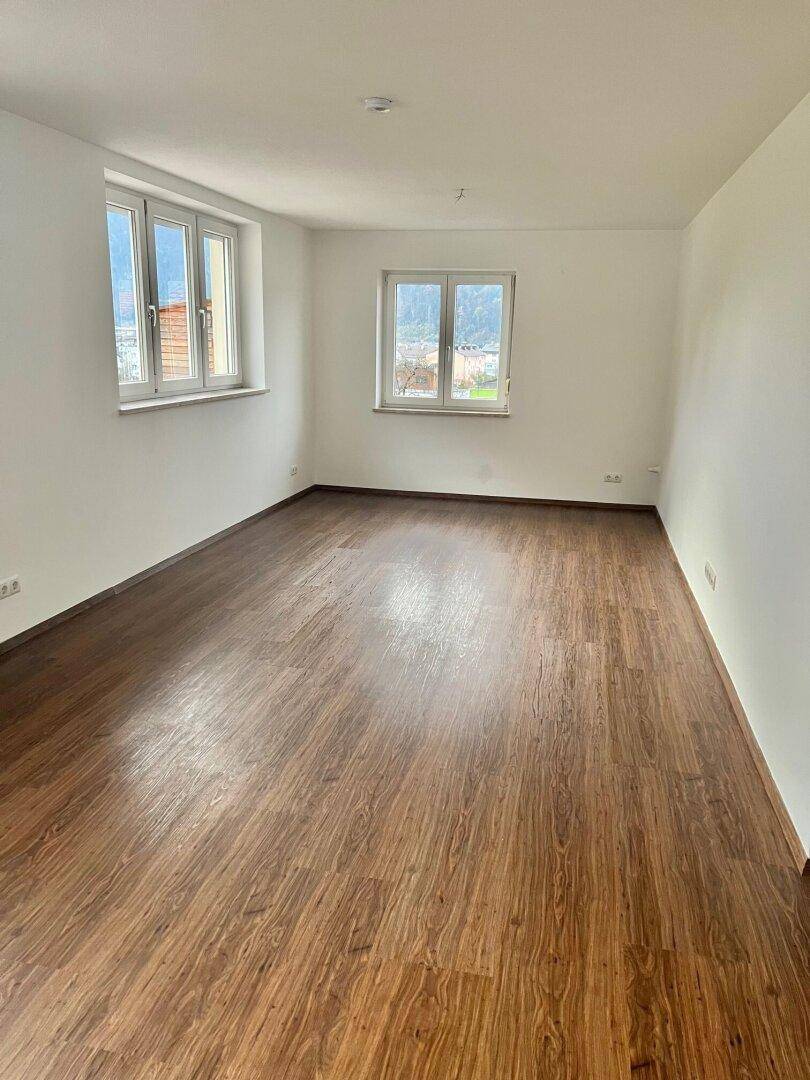 Geräumiges Wohnzimmer mit Holzboden und großen Fenstern, die viel Tageslicht hereinlassen.