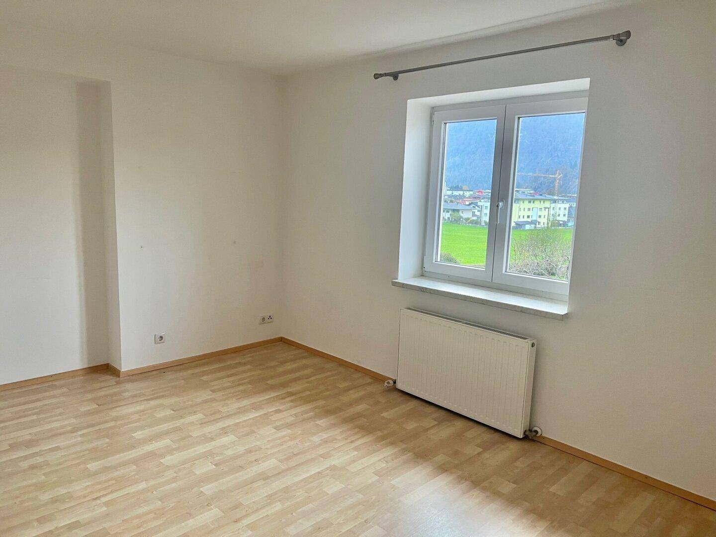 Schlafzimmer mit Holzboden und großem Fenster, das viel Tageslicht und eine grüne Aussicht bietet.