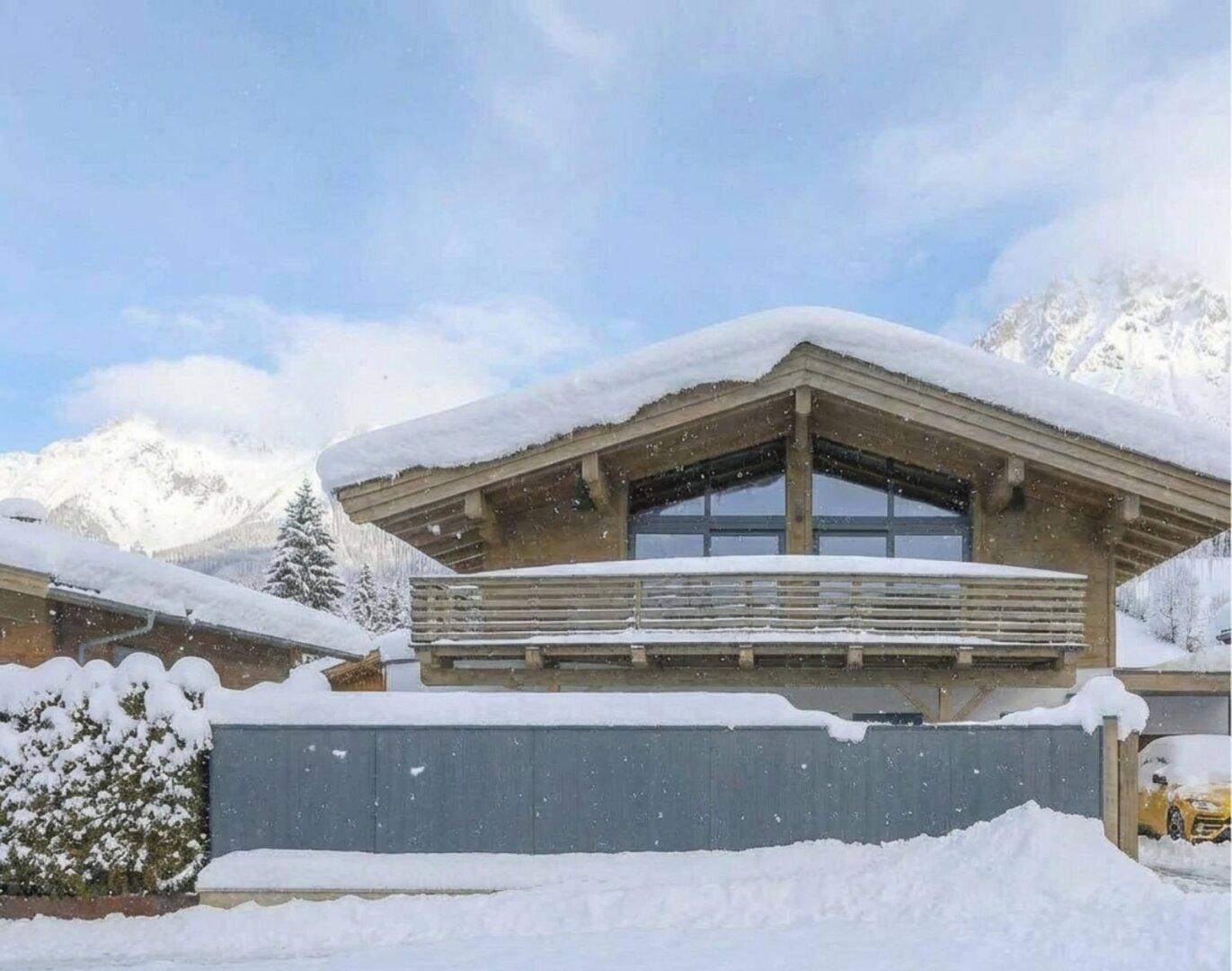 Modernes Chalet mit Holzbalkon und großen Fenstern, umgeben von Schnee und Bergen.