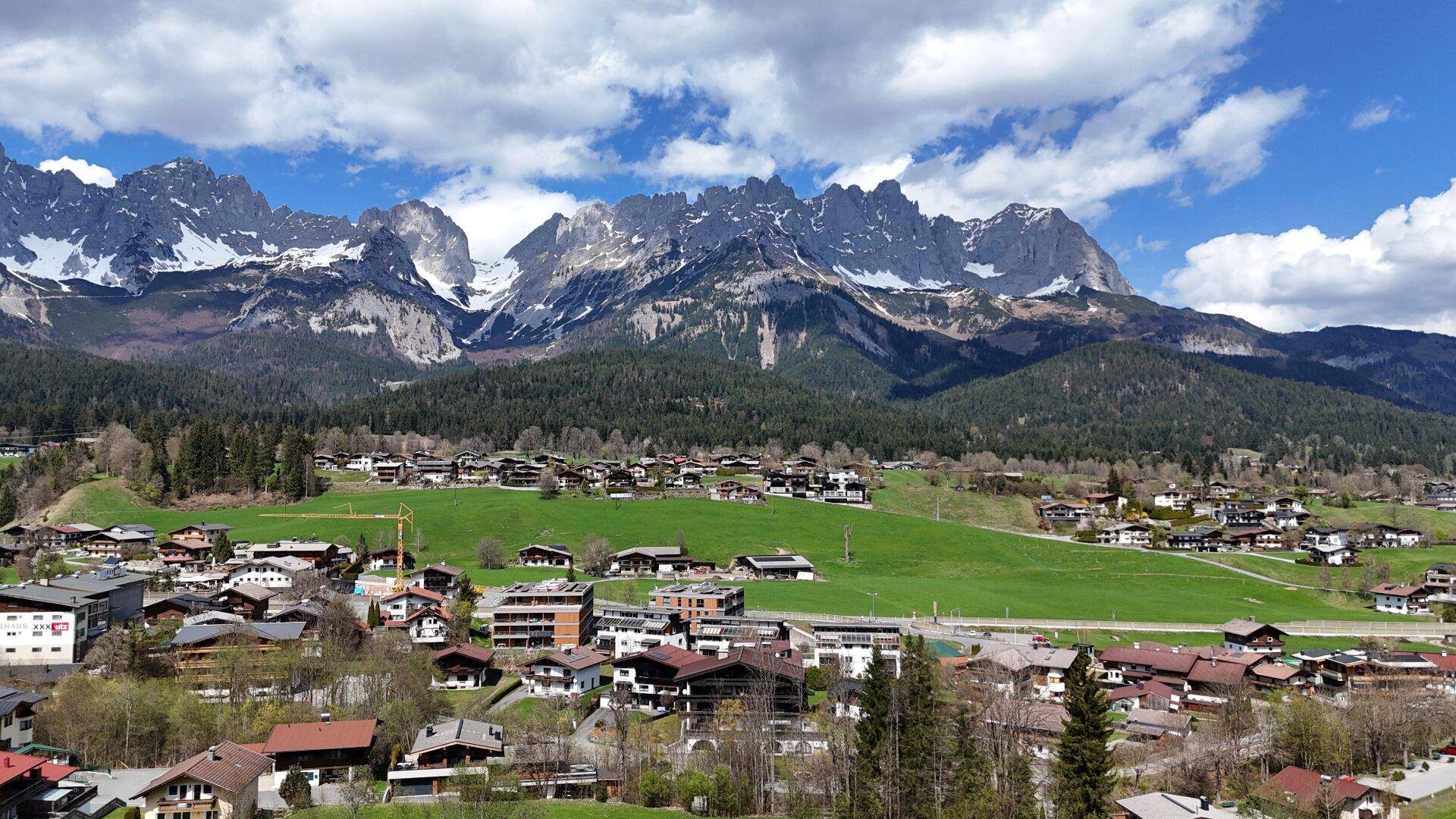 Weitläufige Ansicht eines Bergdorfes mit zahlreichen Häusern und weiten grünen Wiesen vor einer beeindruckenden Bergkulisse.