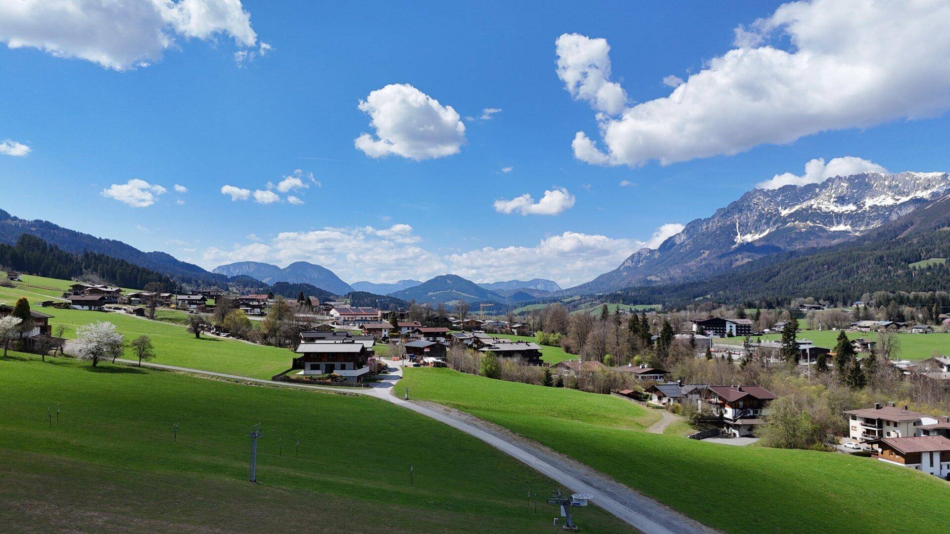 Panoramablick auf ein malerisches Dorf in einem grünen Tal, umgeben von majestätischen Bergen unter blauem Himmel.