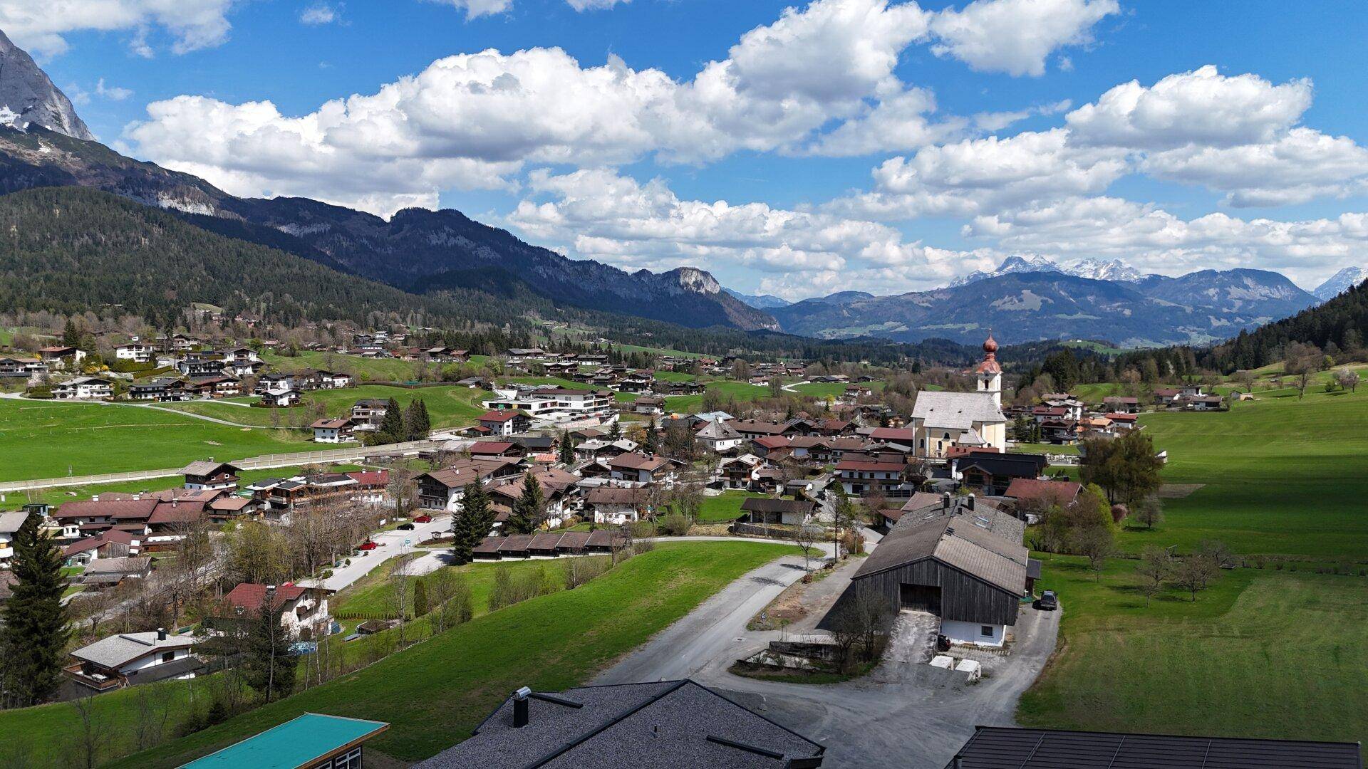 Idyllische Luftaufnahme eines Dorfes mit einer markanten Kirche, eingebettet in grüne Wiesen und umgeben von Bergen.