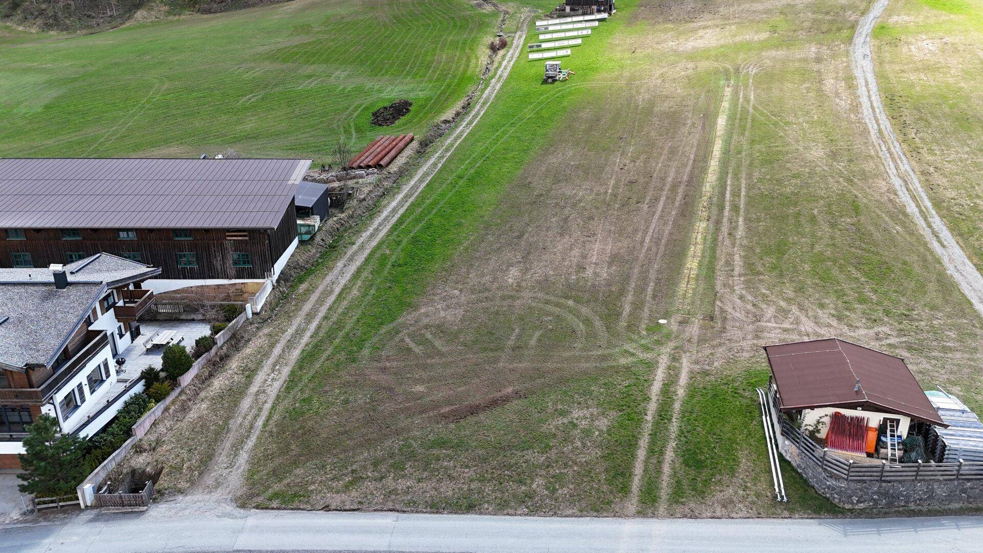 Luftaufnahme eines weitläufigen grünen Feldes mit Fahrspuren, angrenzend an Gebäude und eine ländliche Straße.