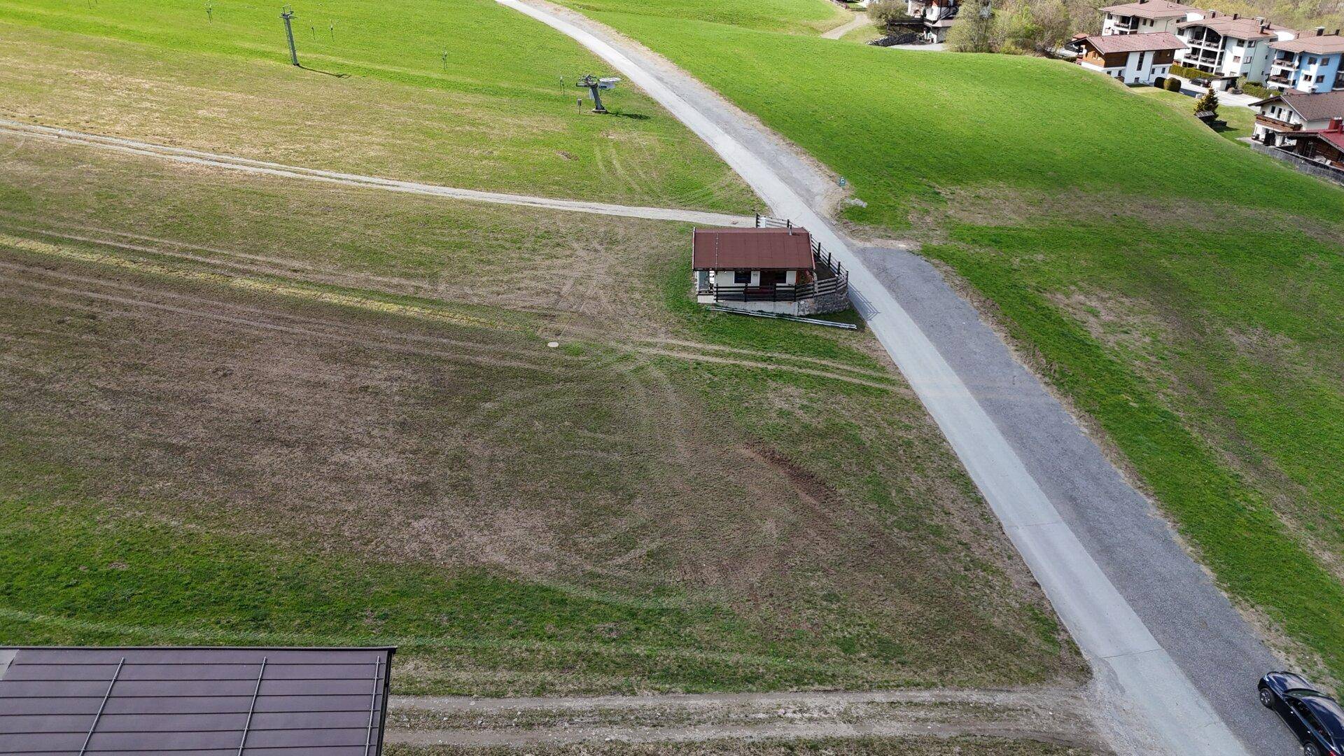 Weitläufige grüne Wiesen mit einer Straße und einem kleinen Gebäude, im Hintergrund sind Skiliftmasten erkennbar.