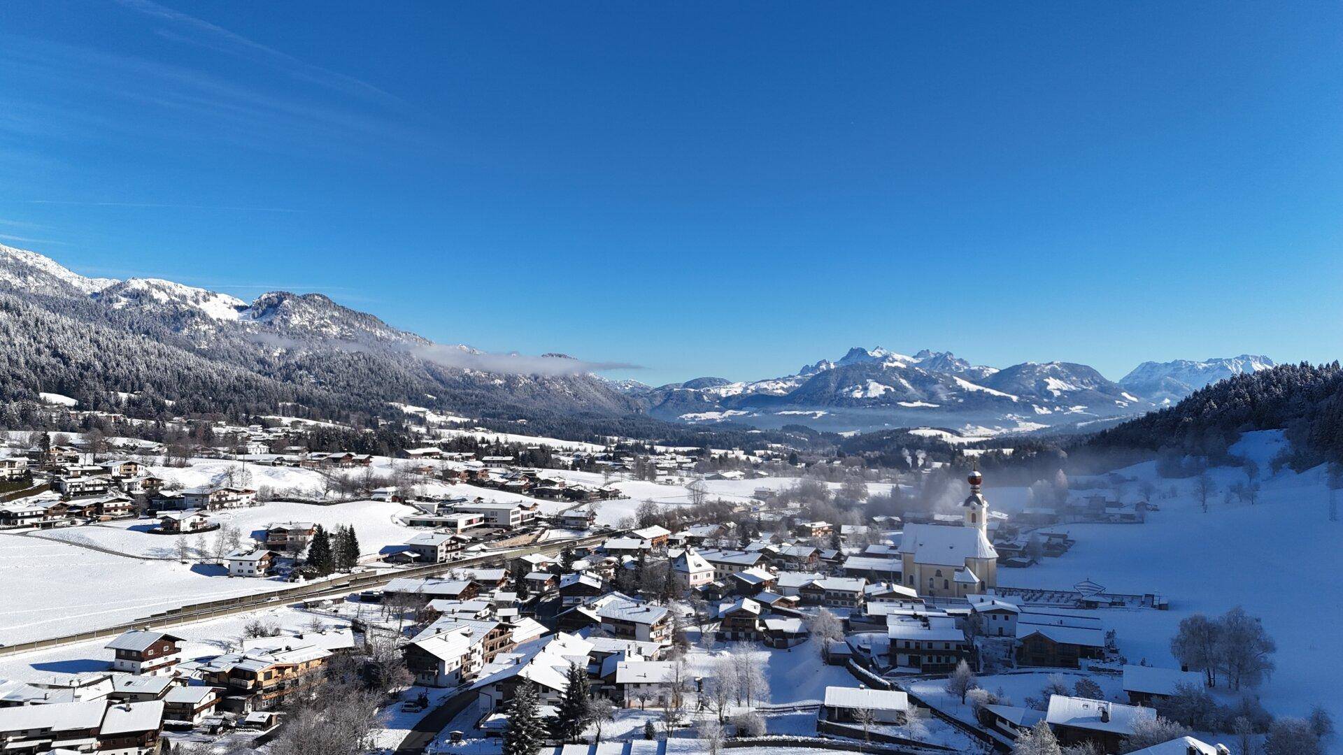 Winterliche Luftaufnahme eines verschneiten Dorfes mit Kirche, umgeben von schneebedeckten Bergen unter klarem Himmel.