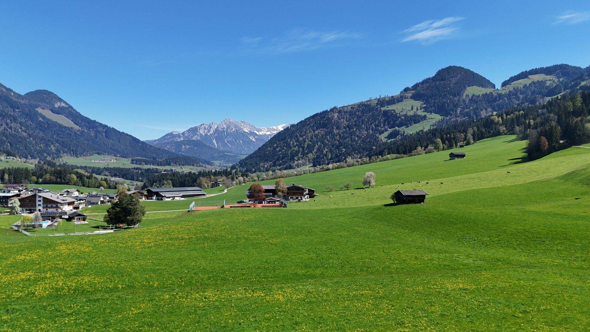 Weitläufiger Panoramablick auf ein Tal mit Dorf, grünen Feldern und schneebedeckten Bergen.