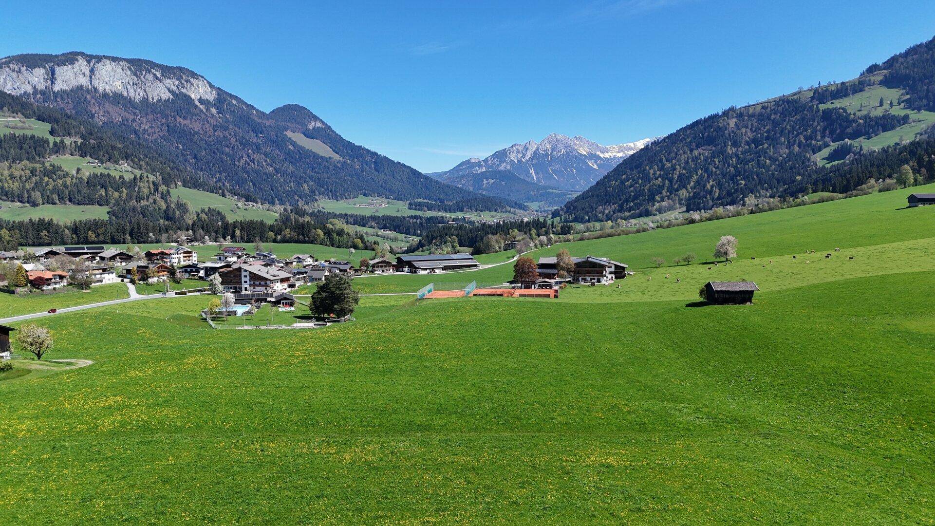 Weitläufiger Panoramablick auf ein Tal mit Dorf, grünen Feldern und schneebedeckten Bergen im Hintergrund.
