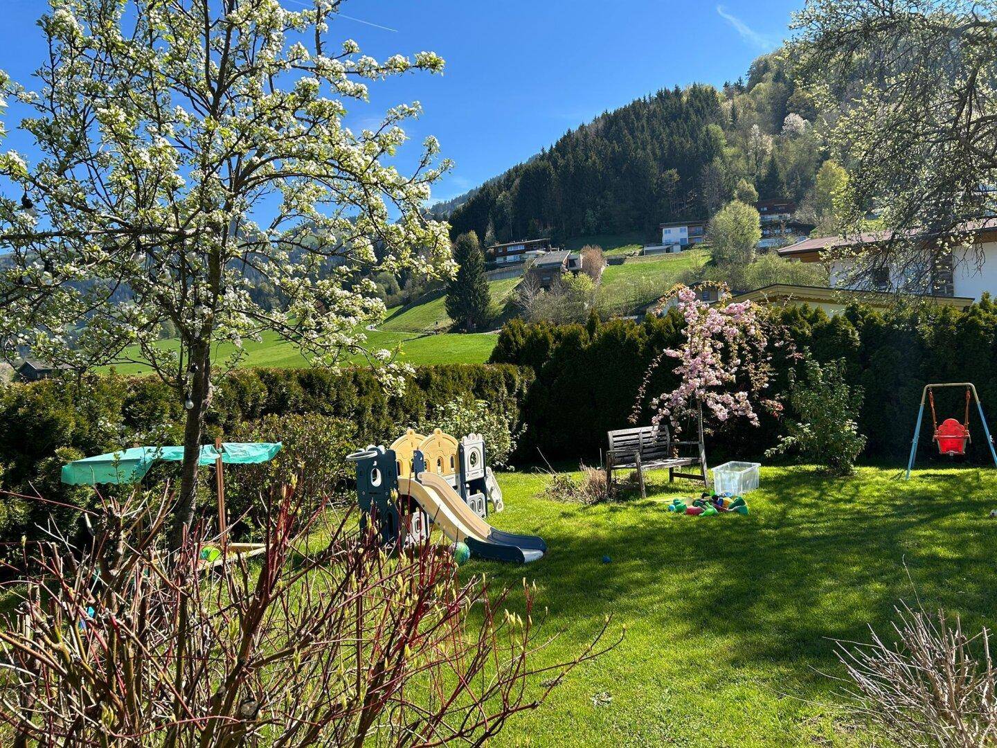 Familienfreundlicher Garten mit Spielplatz, Sitzbank und Blick auf grüne Hügel und Häuser.