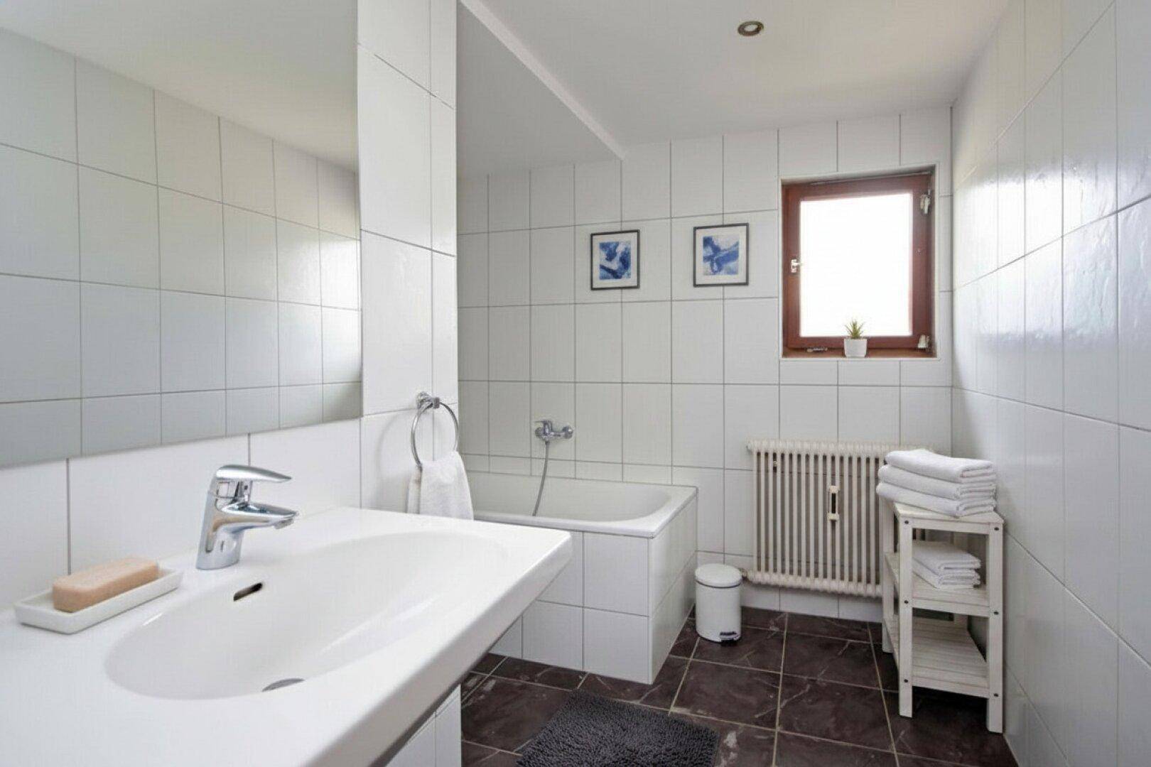 Helles Badezimmer mit weißen Fliesen, Badewanne, Einzelwaschbecken und Fenster.