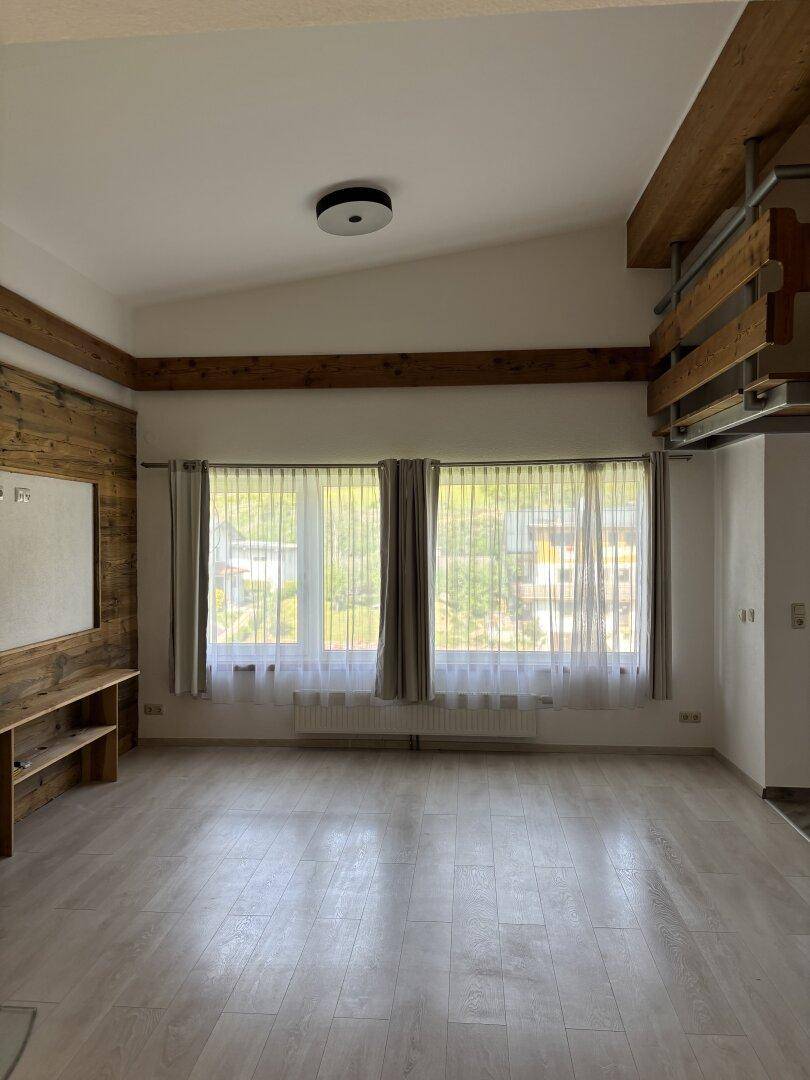 Helles Wohnzimmer mit großen Fenstern, sichtbaren Holzbalken und hellem Laminatboden.