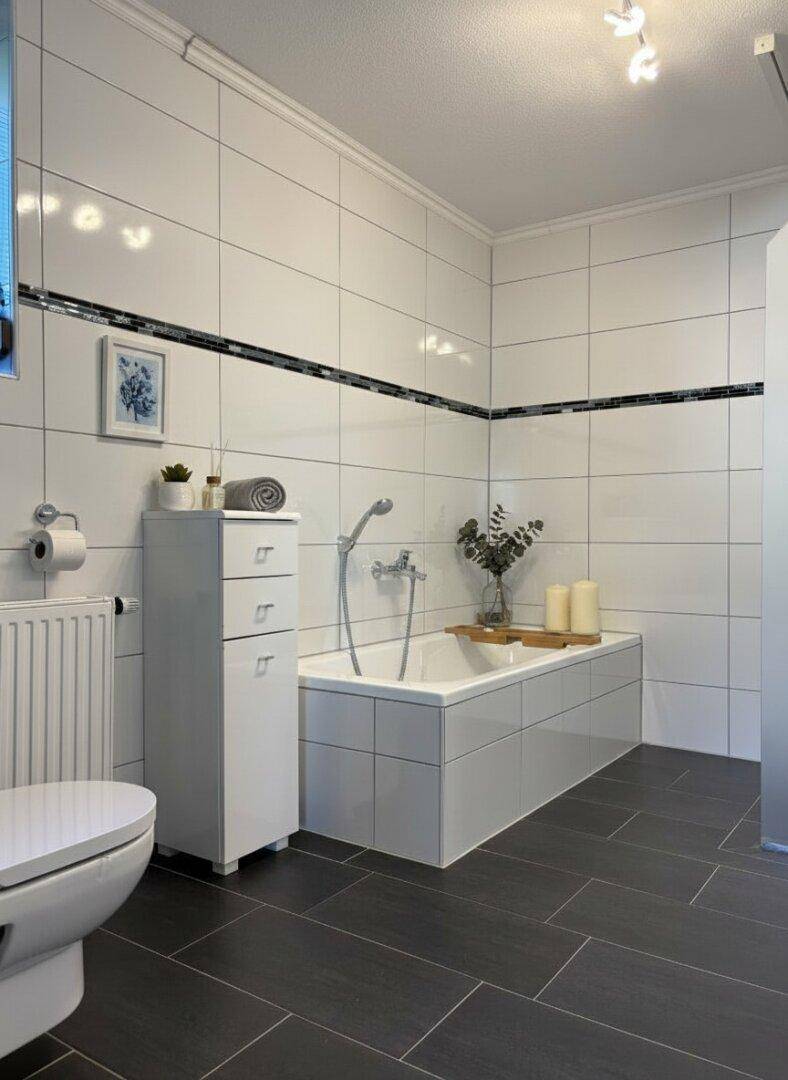 Badezimmer mit Badewanne, Toilette und weißen Fliesen mit schwarzem Dekorstreifen.