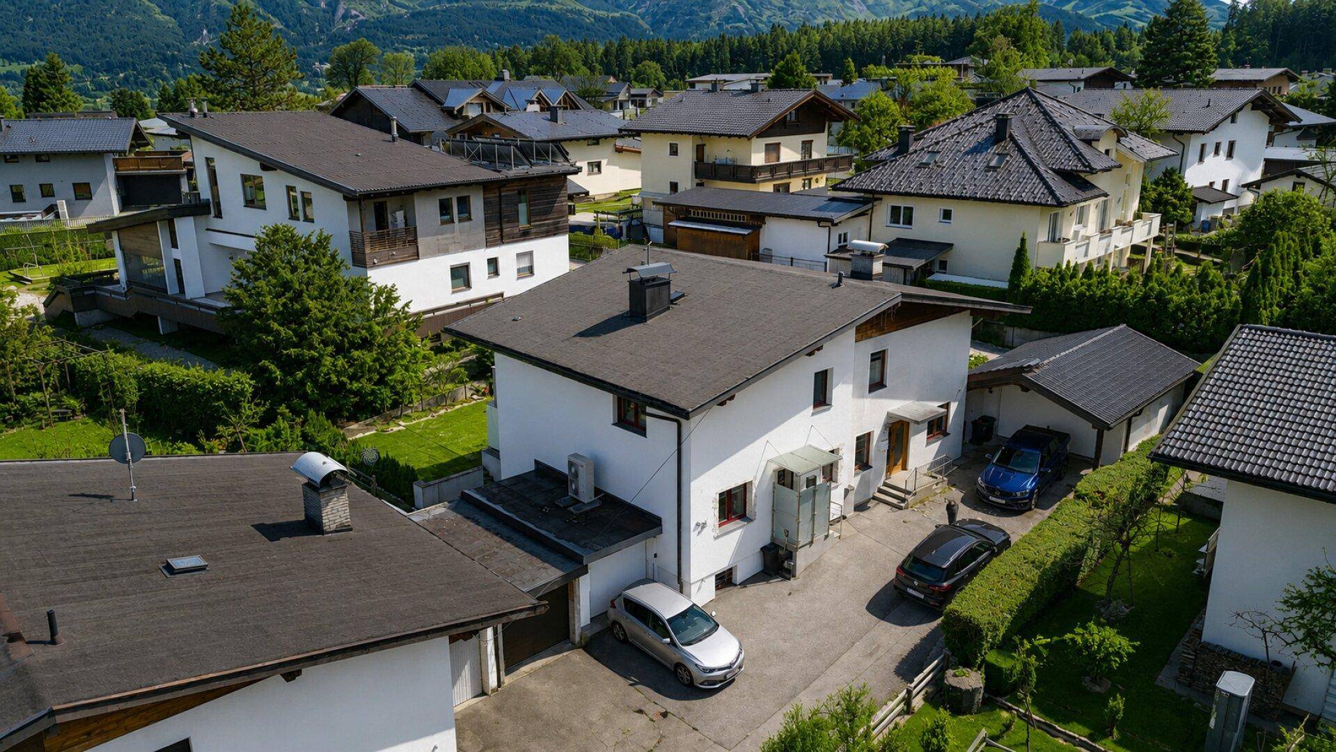 Luftaufnahme des Hauses mit Einfahrt, parkenden Autos und Blick auf die umliegende Wohngegend und Berge.