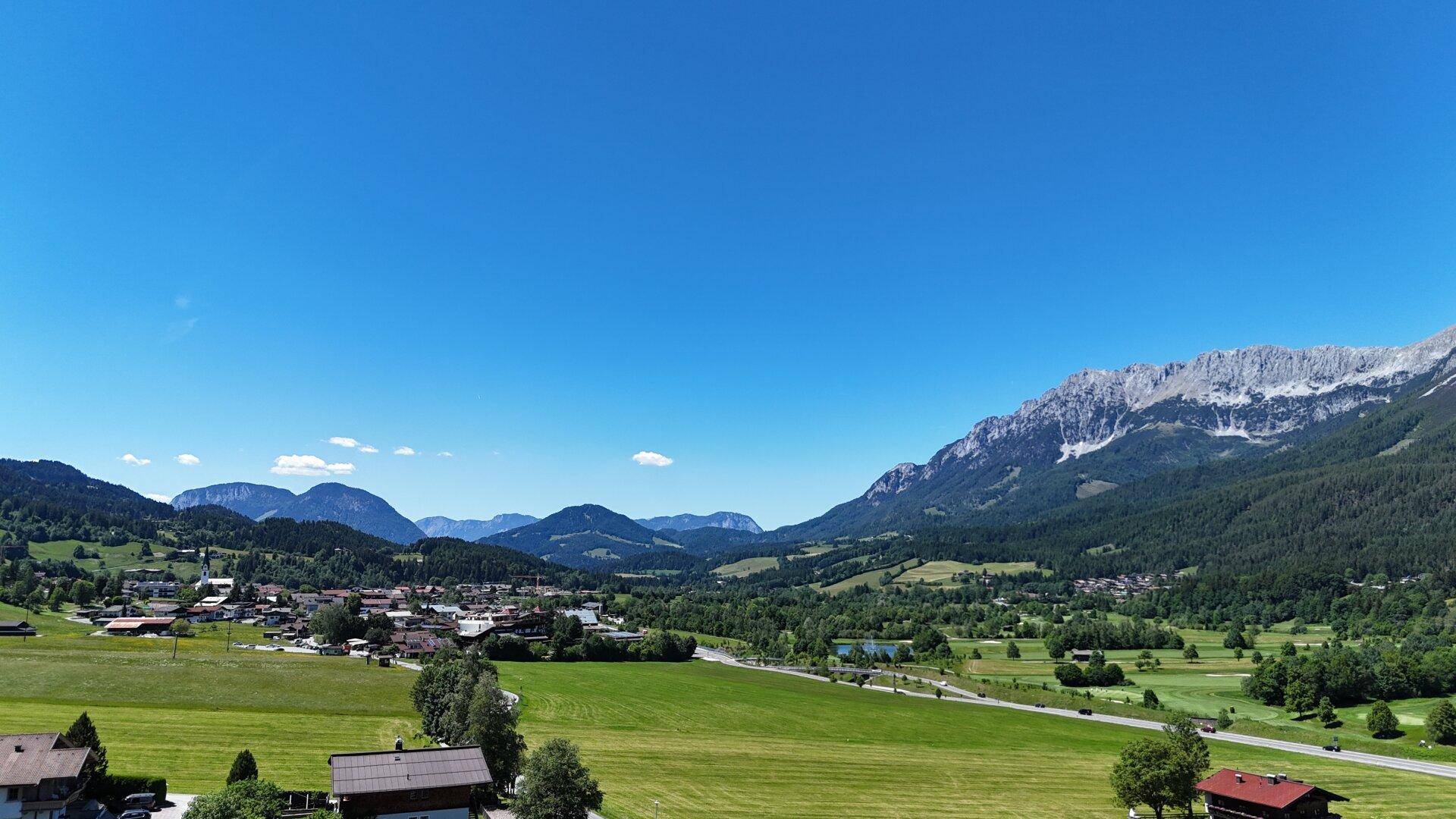 Weitläufiger Blick über ein grünes Tal mit einem Dorf und einer Bergkette am Horizont unter blauem Himmel.