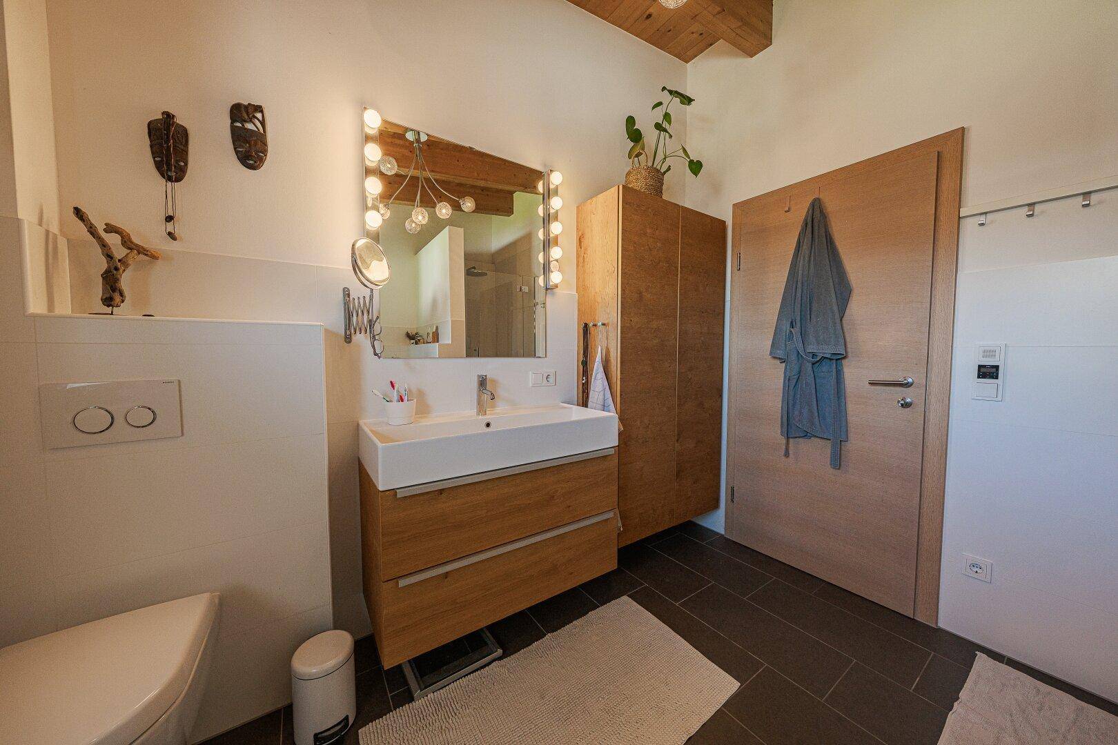 Modernes Badezimmer mit Holzwaschtisch, beleuchtetem Spiegel und dunklen Fliesen.