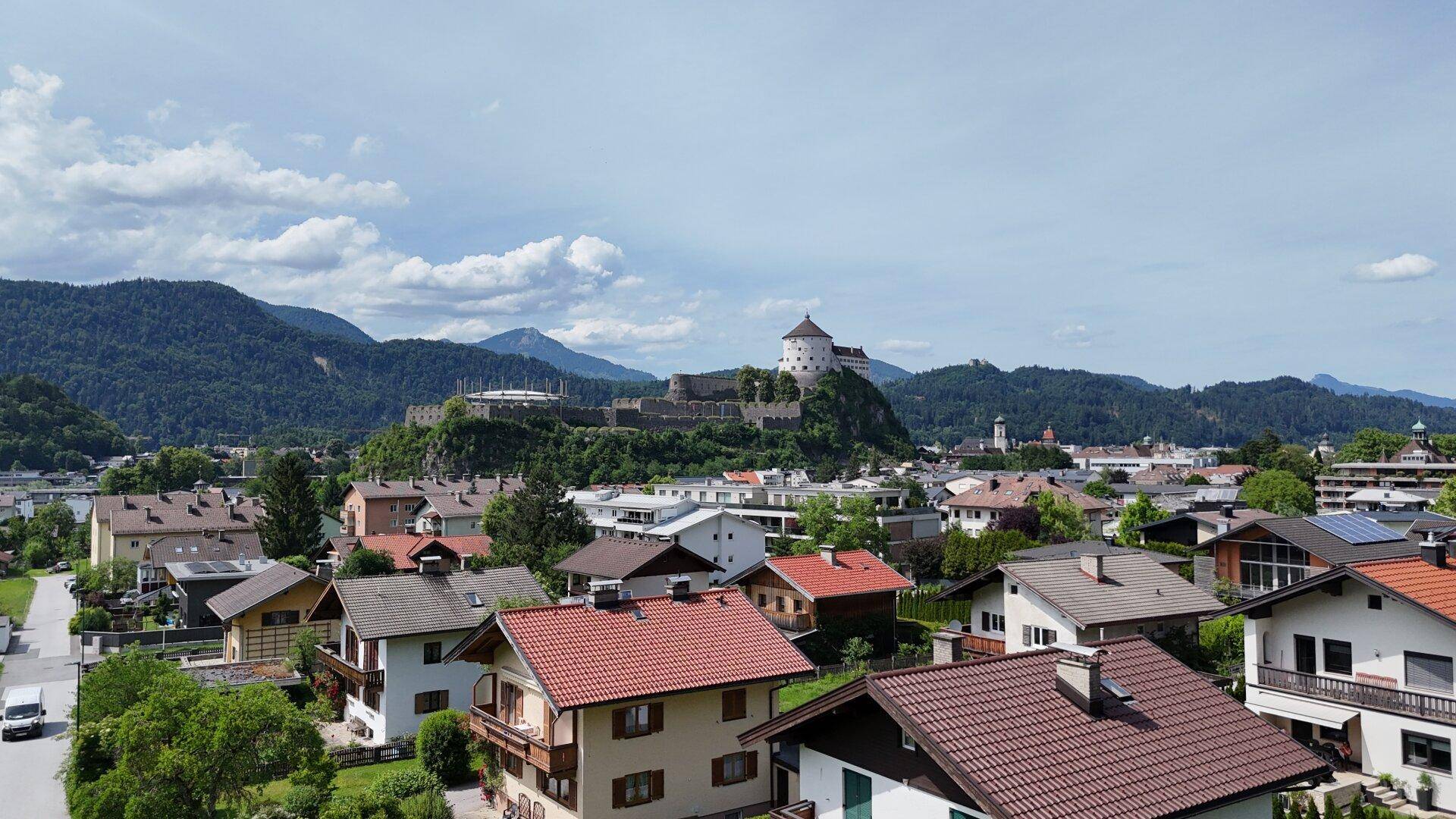 Panoramablick über eine Stadt mit historischen Gebäuden, einer Burg auf einem Hügel und umliegenden Bergen.