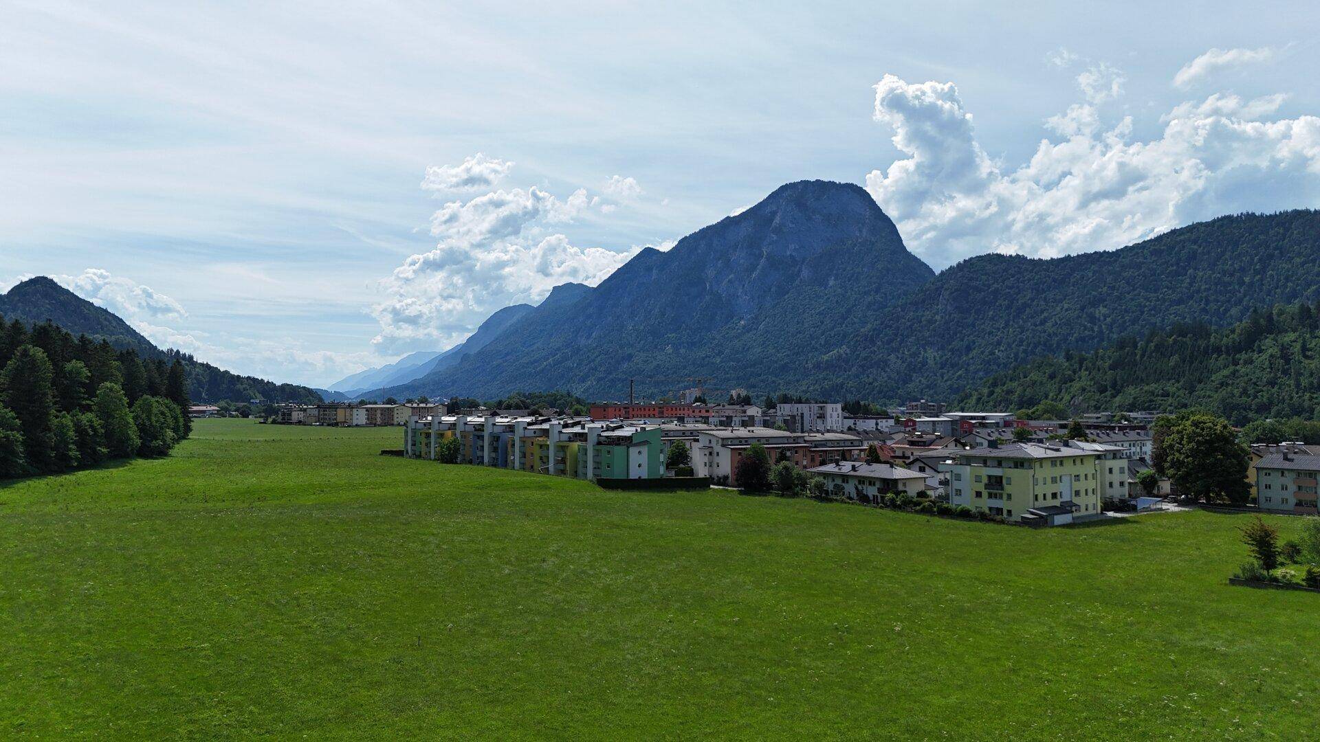 Weitläufige grüne Landschaft mit Wohngebäuden am Horizont und imposanten Bergketten im Hintergrund.