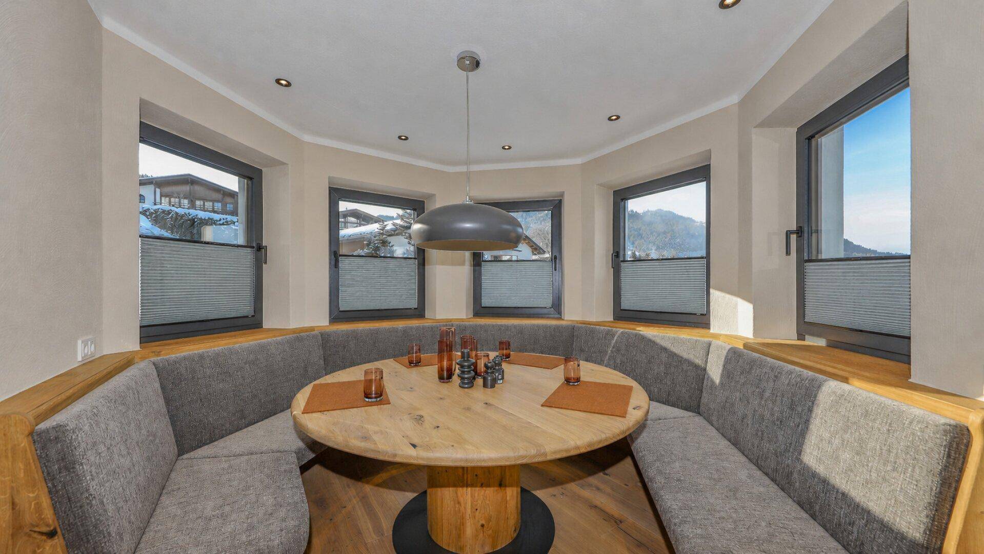 Gemütlicher Essbereich mit runder Holztafel und eingebauter Sitzbank, umgeben von Fenstern mit Bergblick.