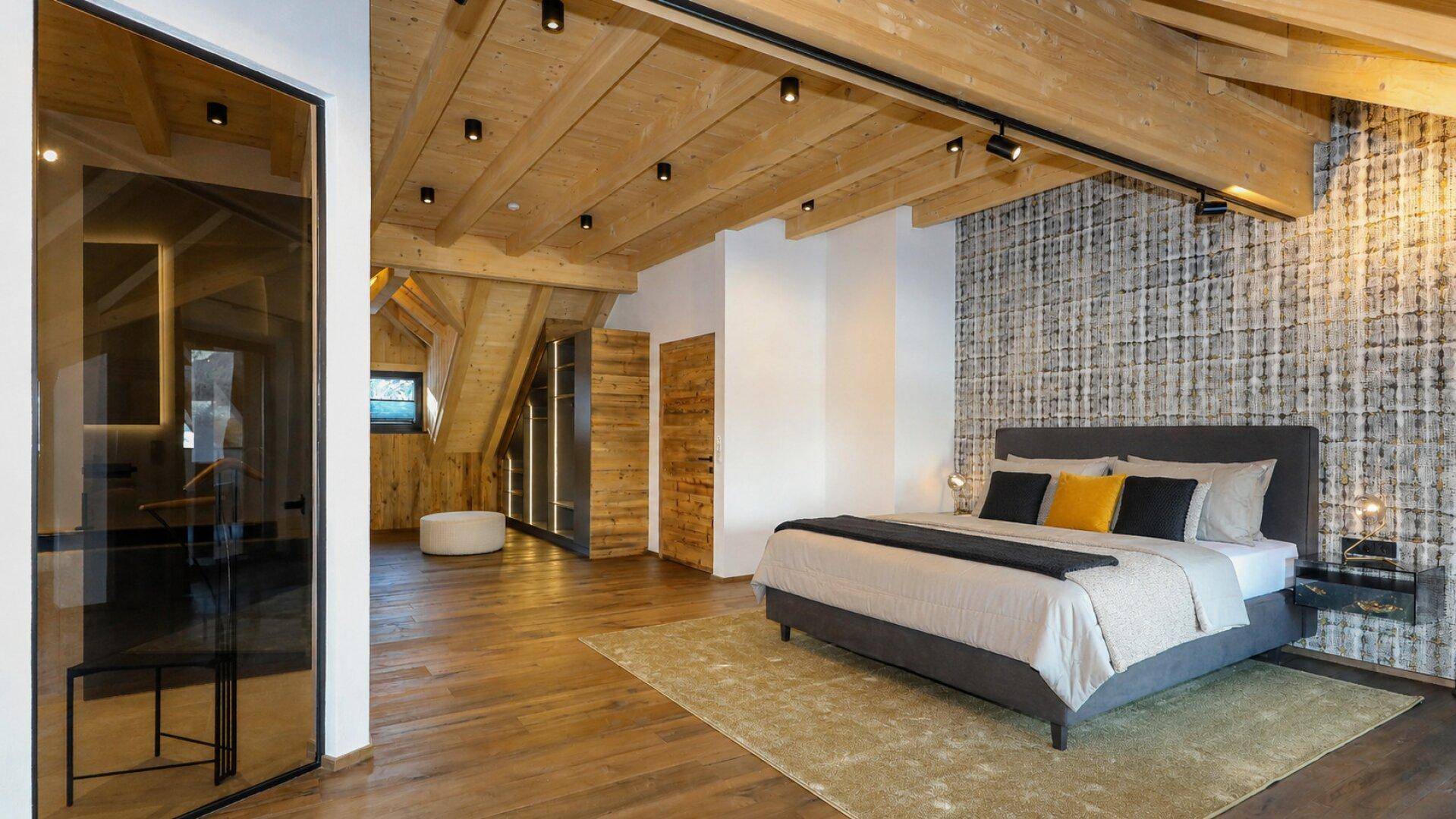 Geräumiges Schlafzimmer mit Holzbalkendecke, Akzentwand und Zugang zu einem modernen Badezimmer.