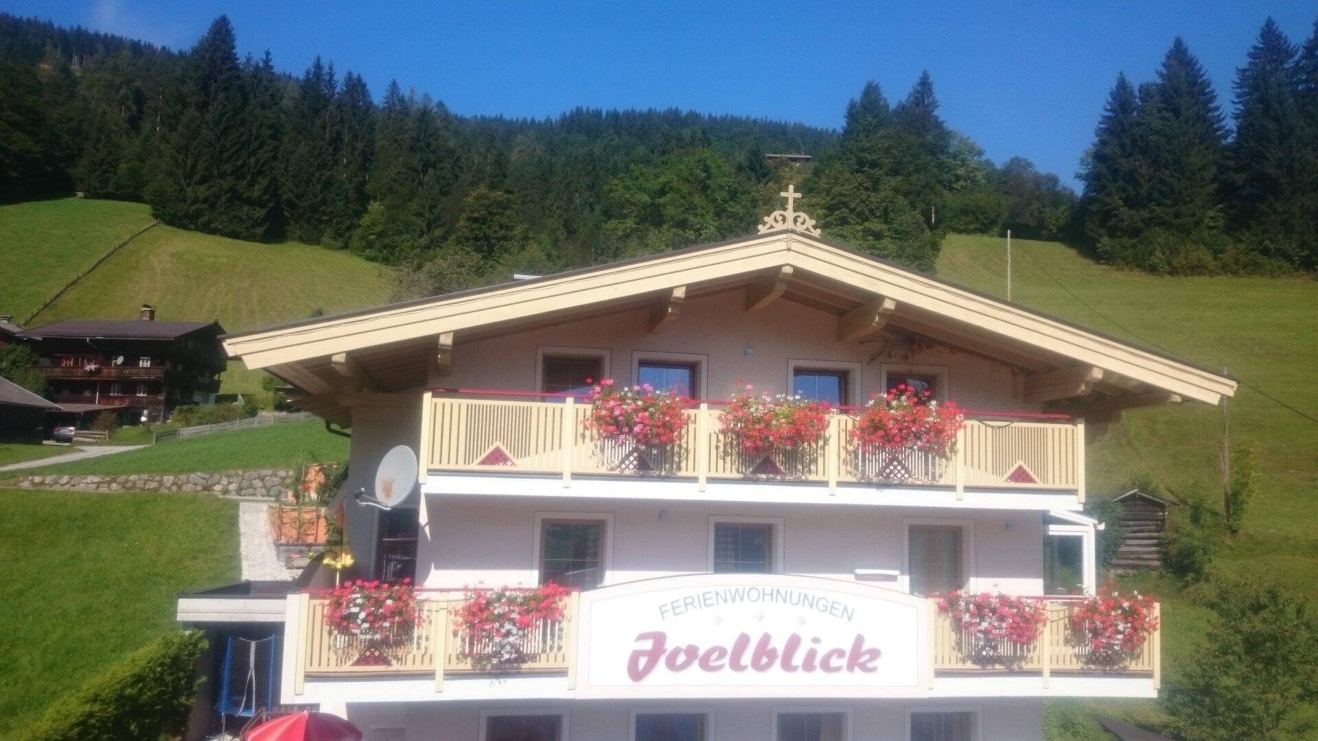 Traditionelles alpines Gebäude mit blühenden Balkonen und bewaldetem Berghintergrund.