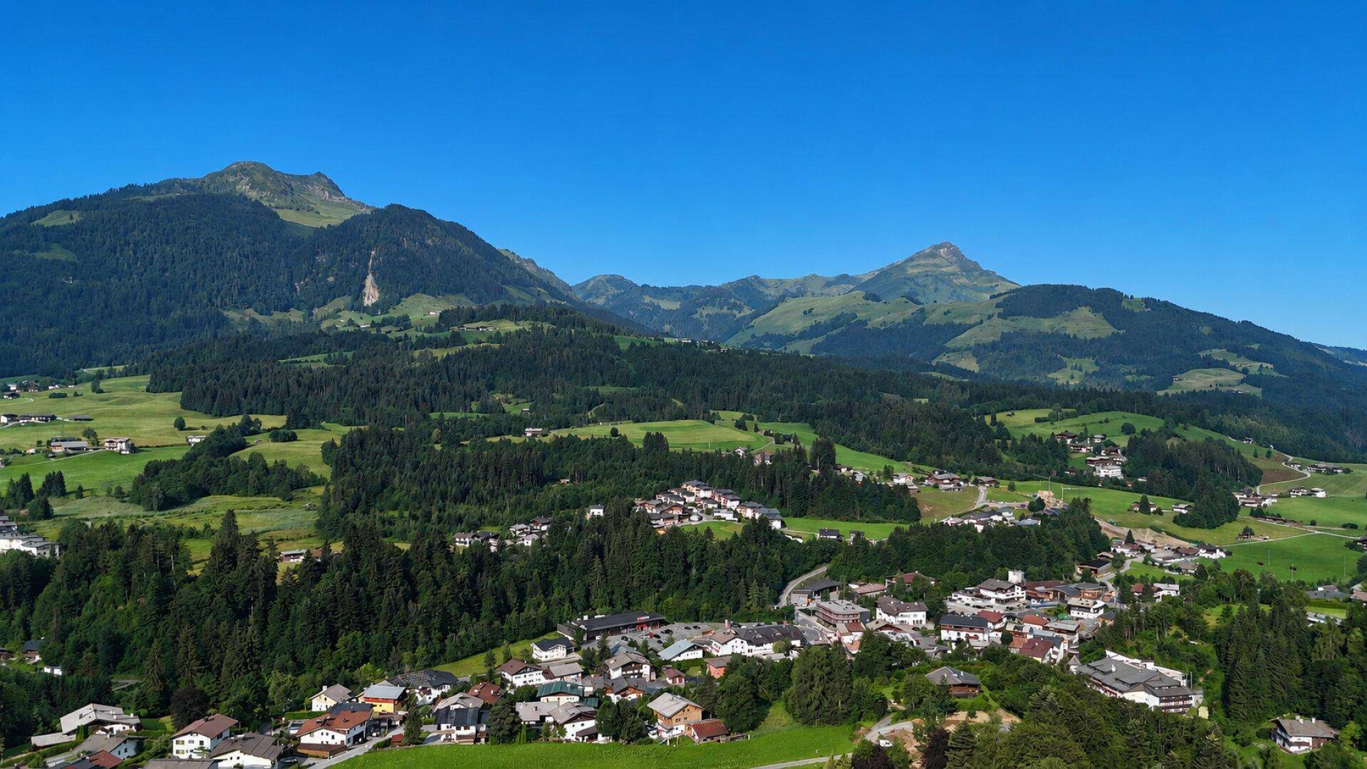 Weitläufiger Panoramablick auf ein idyllisches Dorf, umgeben von bewaldeten Bergen und grünen Wiesen.