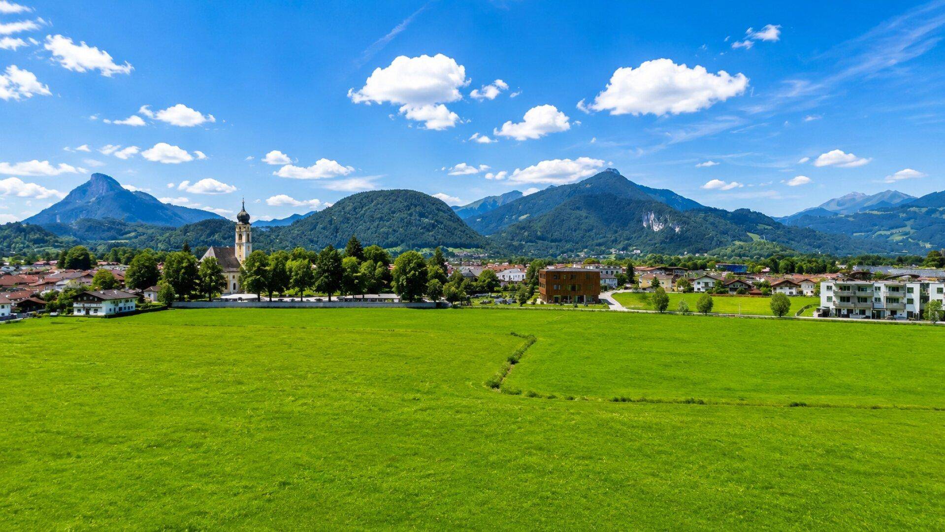Weitläufige grüne Landschaft mit einem charmanten Dorf, einer Kirche und Bergen im Hintergrund.