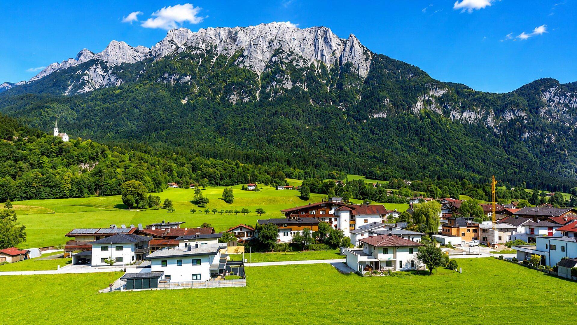 Malerische Landschaft mit einem Dorf im Tal, umgeben von grünen Wiesen und imposanten Bergen.