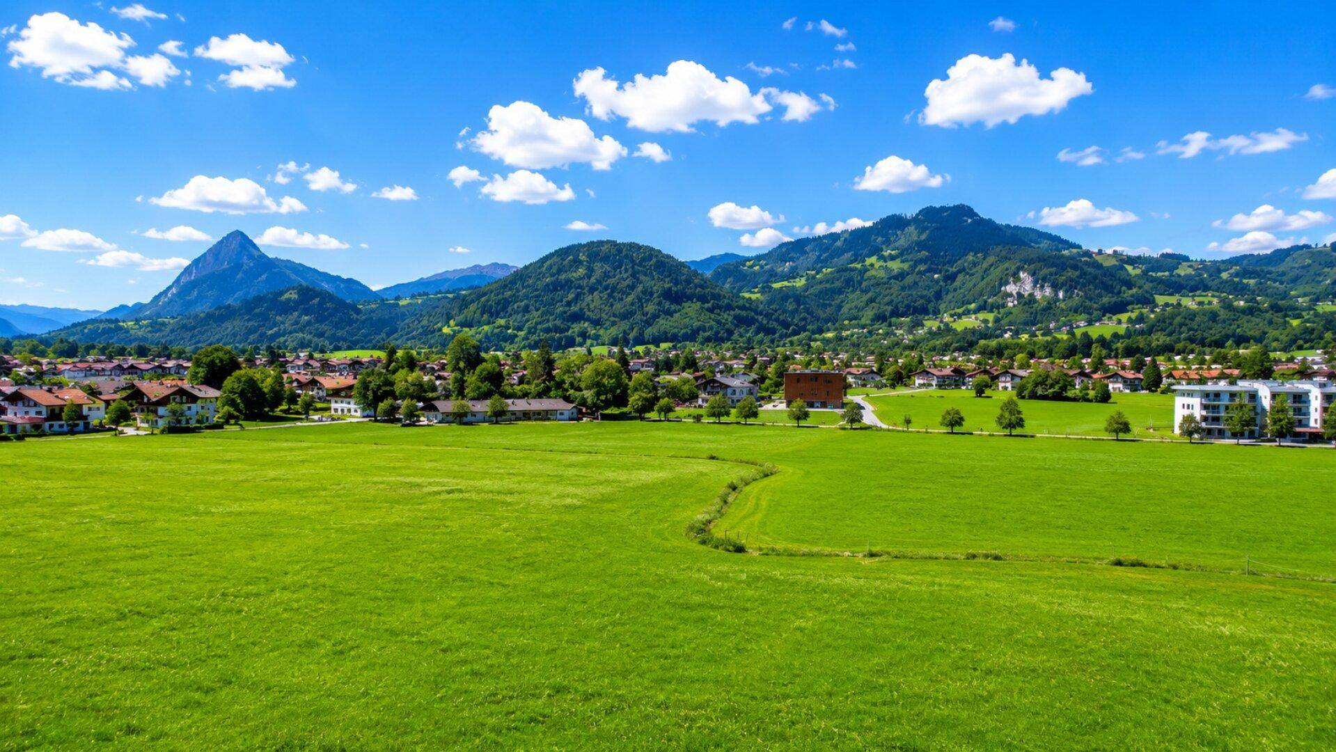 Panoramablick über grüne Felder auf eine malerische Stadt mit Bergen im Hintergrund.