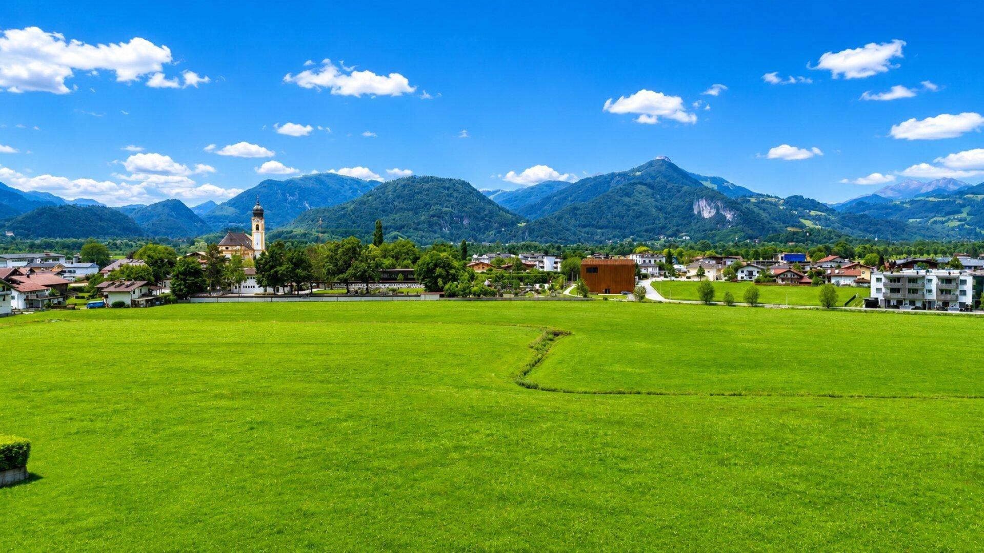Weitläufige grüne Landschaft mit einer charmanten Stadt und imposanten Bergen unter blauem Himmel.