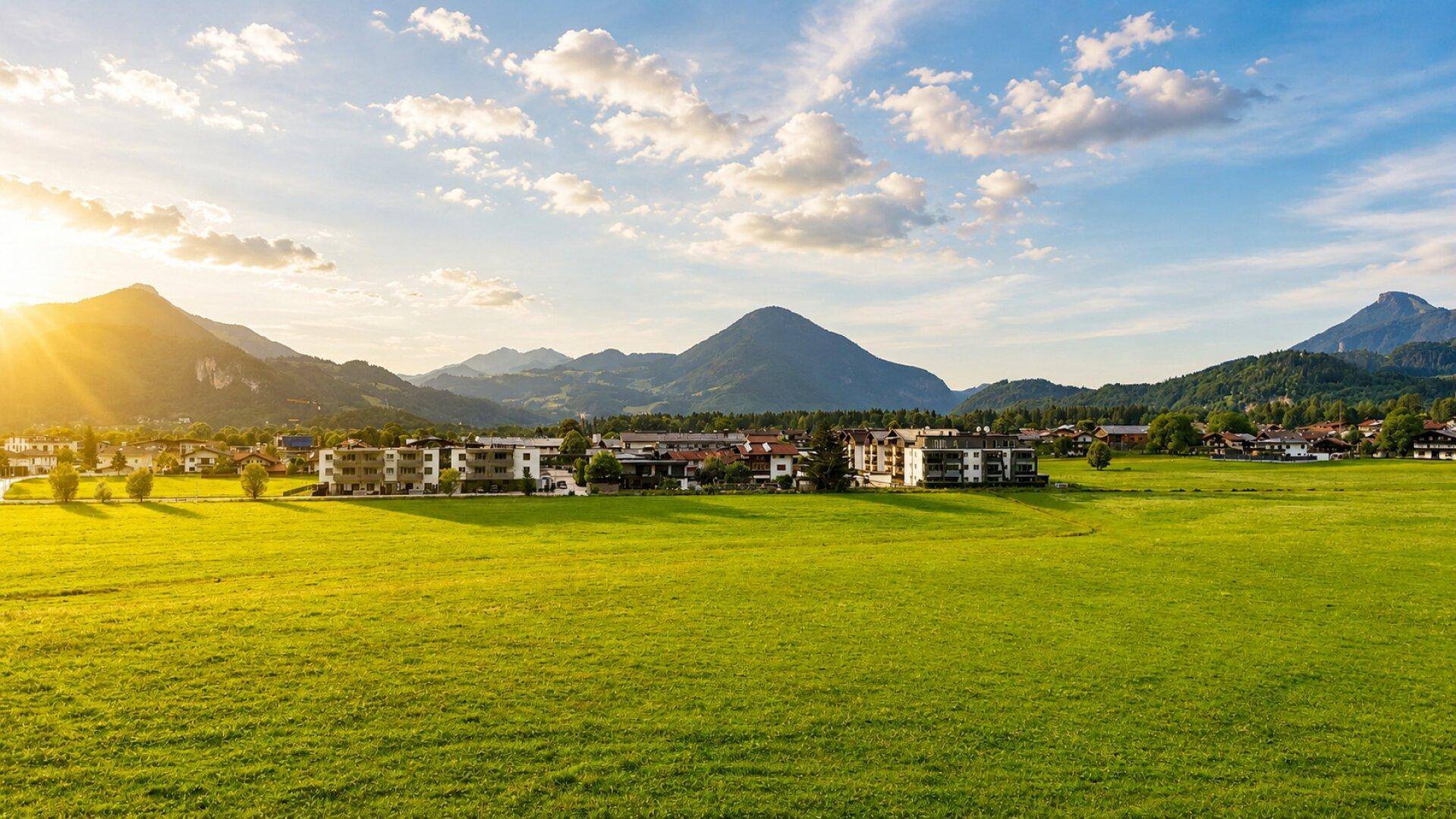 Sonnenuntergang über weiten grünen Feldern mit einer Stadt und majestätischen Bergen am Horizont.