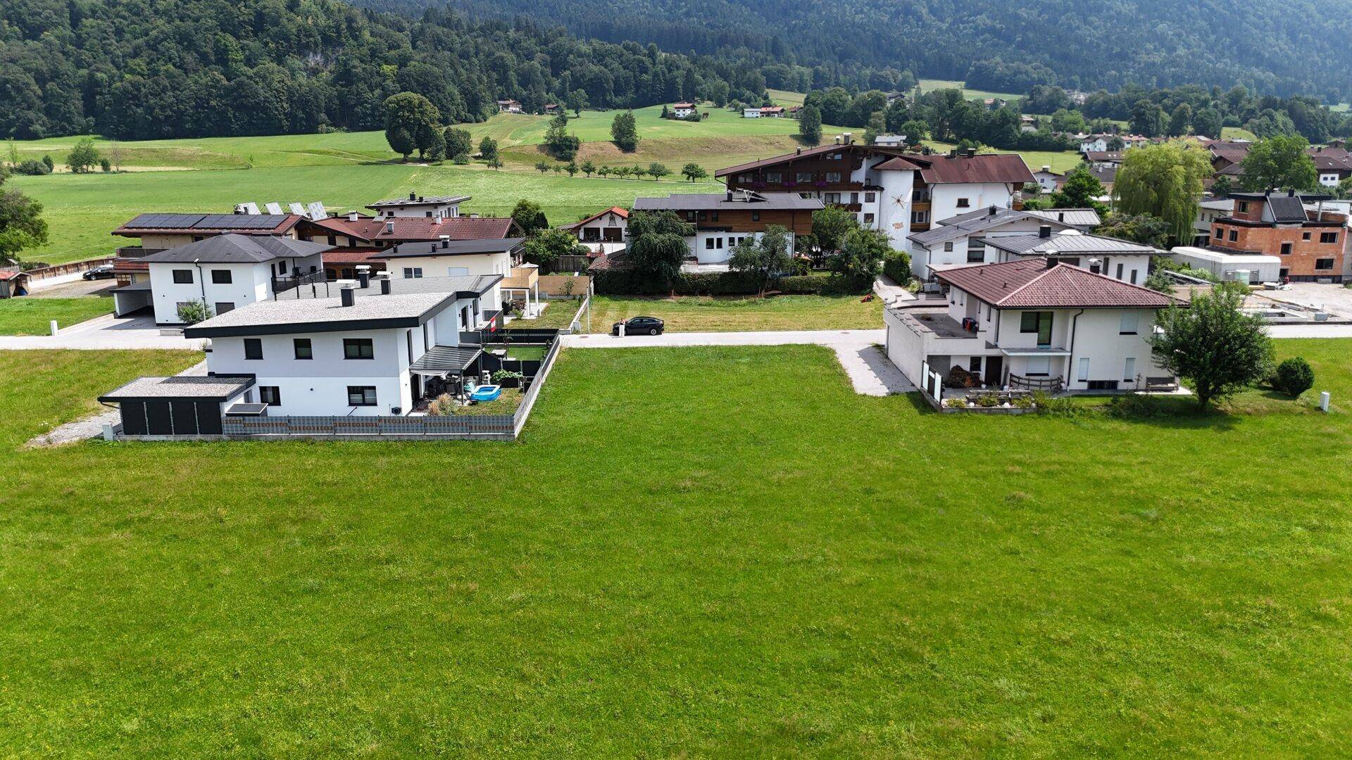 Grüne Wiesenfläche mit mehreren Wohnhäusern und einer Zufahrtsstraße in ländlicher Umgebung.