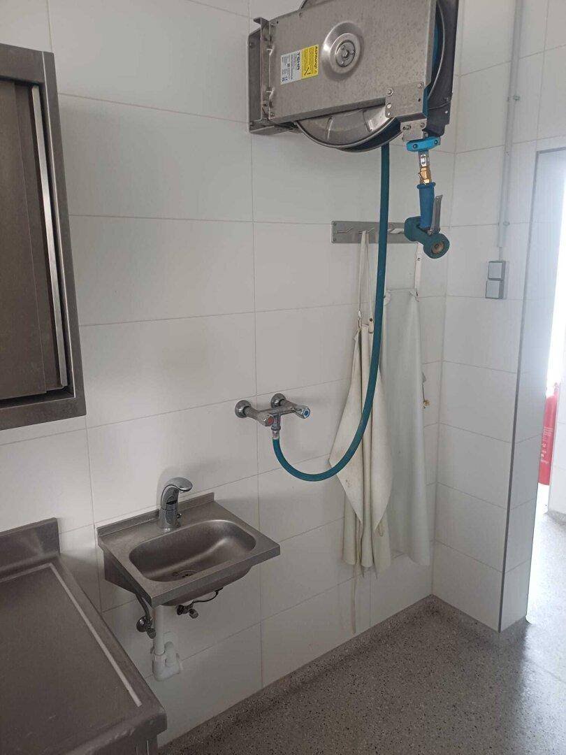 Kleines Edelstahlwaschbecken mit Wandarmatur und Schlauchaufroller in einem Reinigungsbereich.