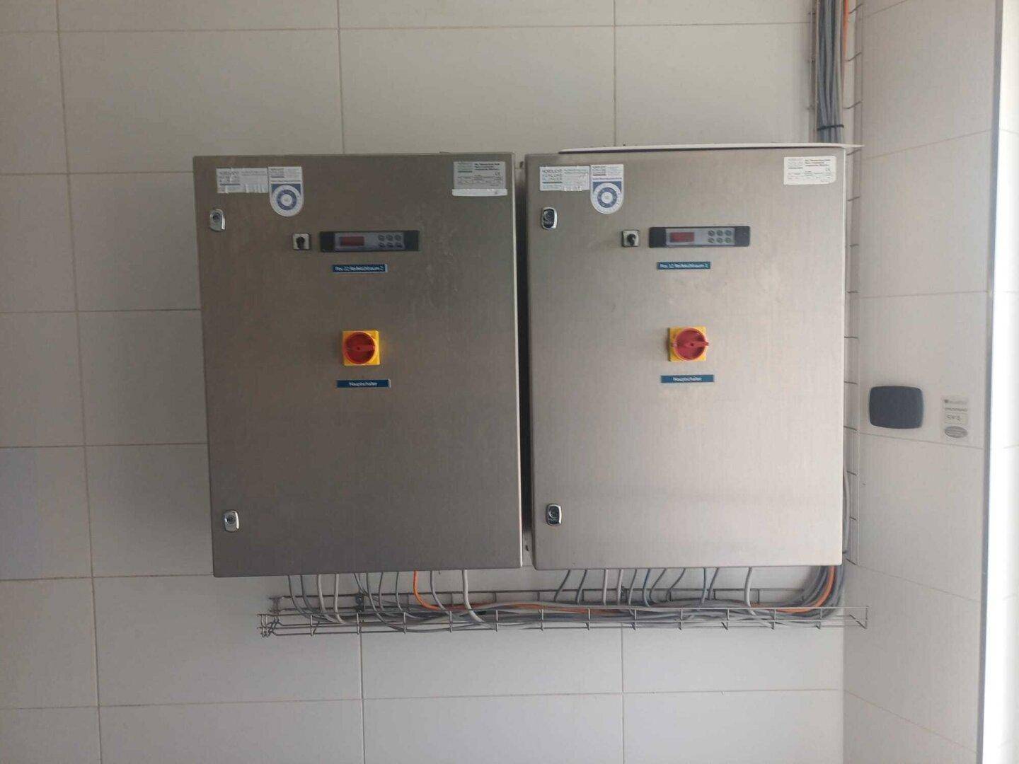 Zwei elektrische Schalttafeln mit Bedienelementen und Verkabelung an einer gefliesten Wand.
