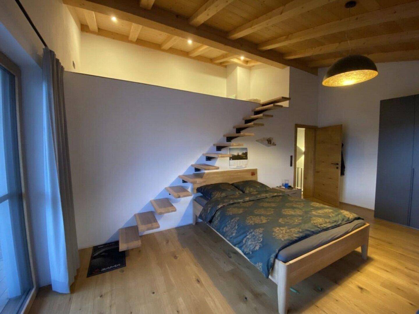 Helles Schlafzimmer mit Holzbalkendecke, schwebender Treppe zum Loft und großem Fenster.
