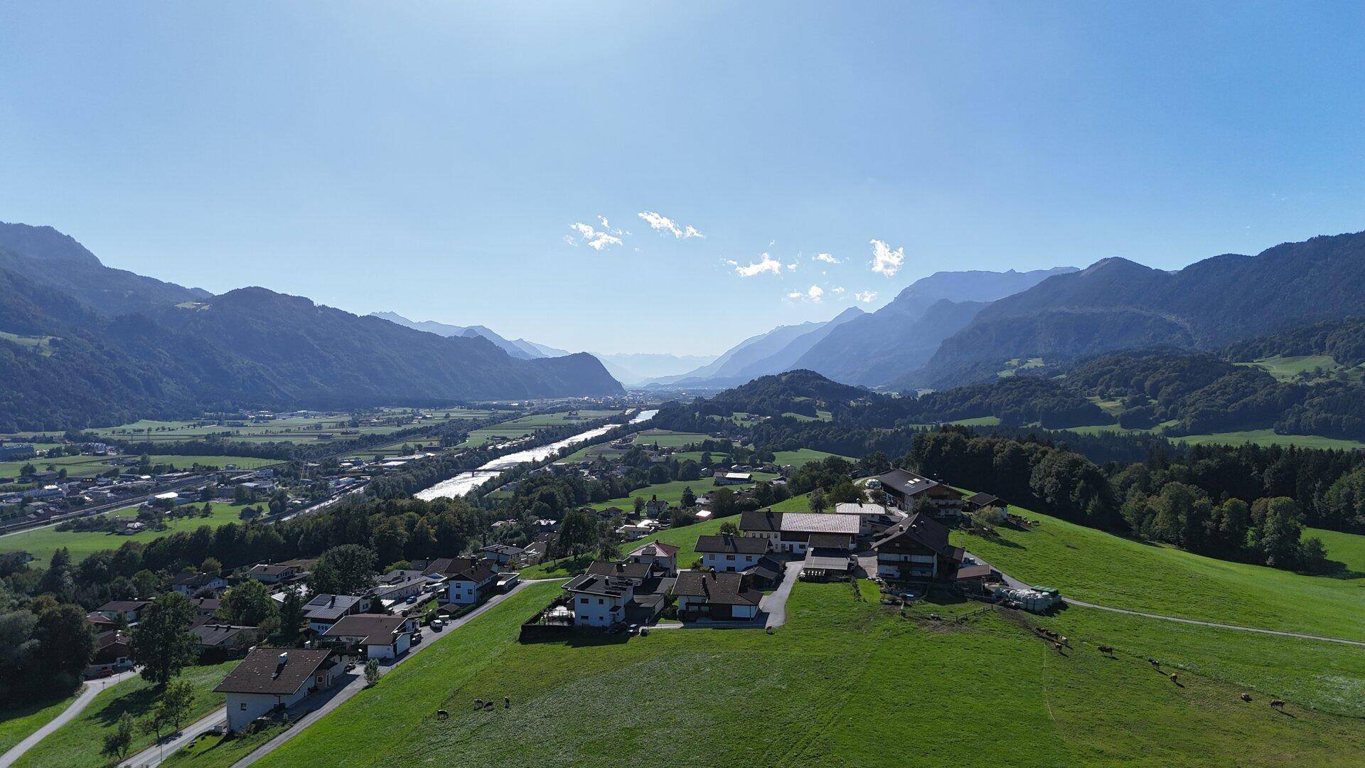 Dorf auf einem Hügel mit Blick auf ein Flusstal und eine beeindruckende Bergkette unter blauem Himmel.