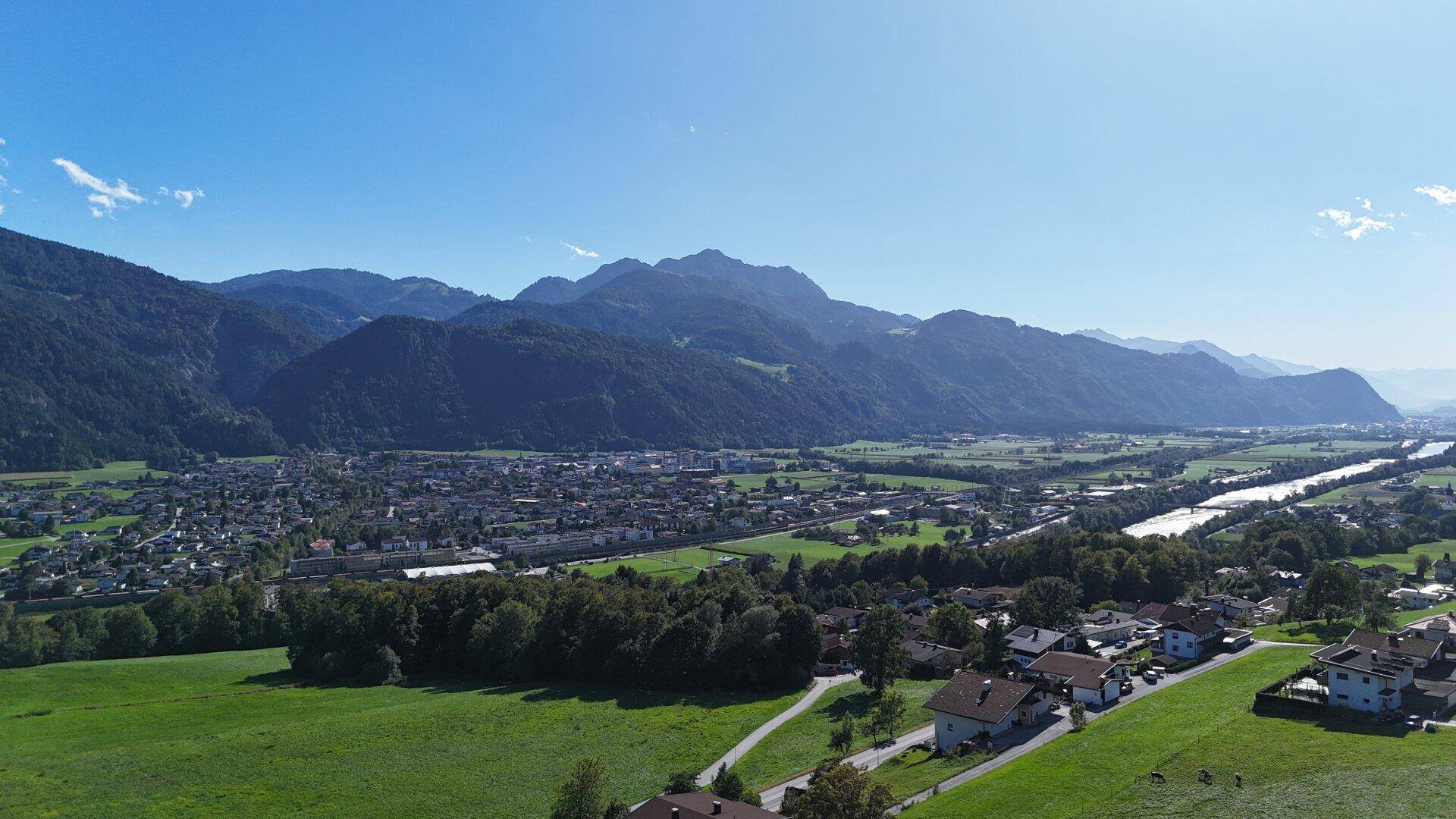 Luftaufnahme einer Stadt im Tal, umgeben von bewaldeten Bergen und einem Fluss, unter klarem Himmel.
