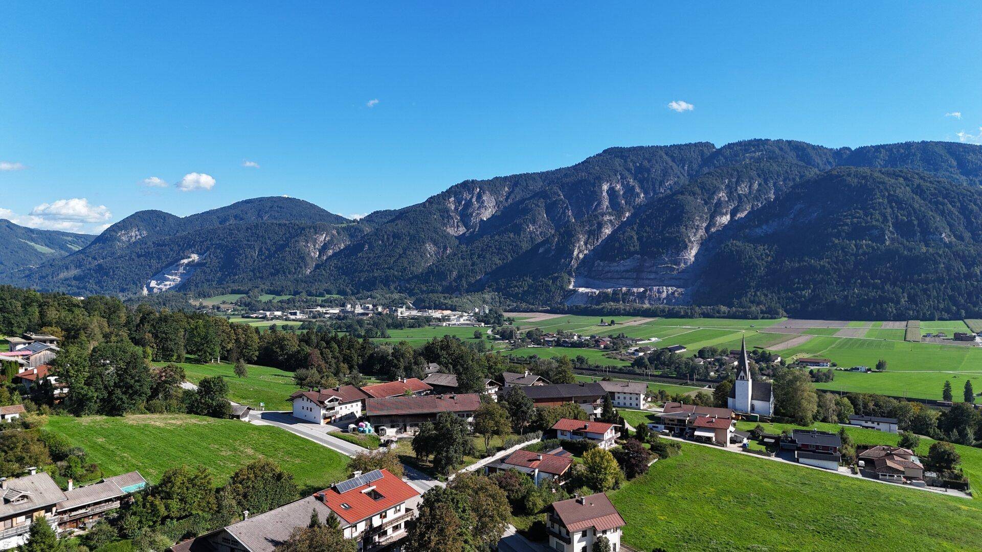 Ländliche Szenerie mit einem Dorf, einer Kirche, weiten Feldern und majestätischen Bergen im Hintergrund.