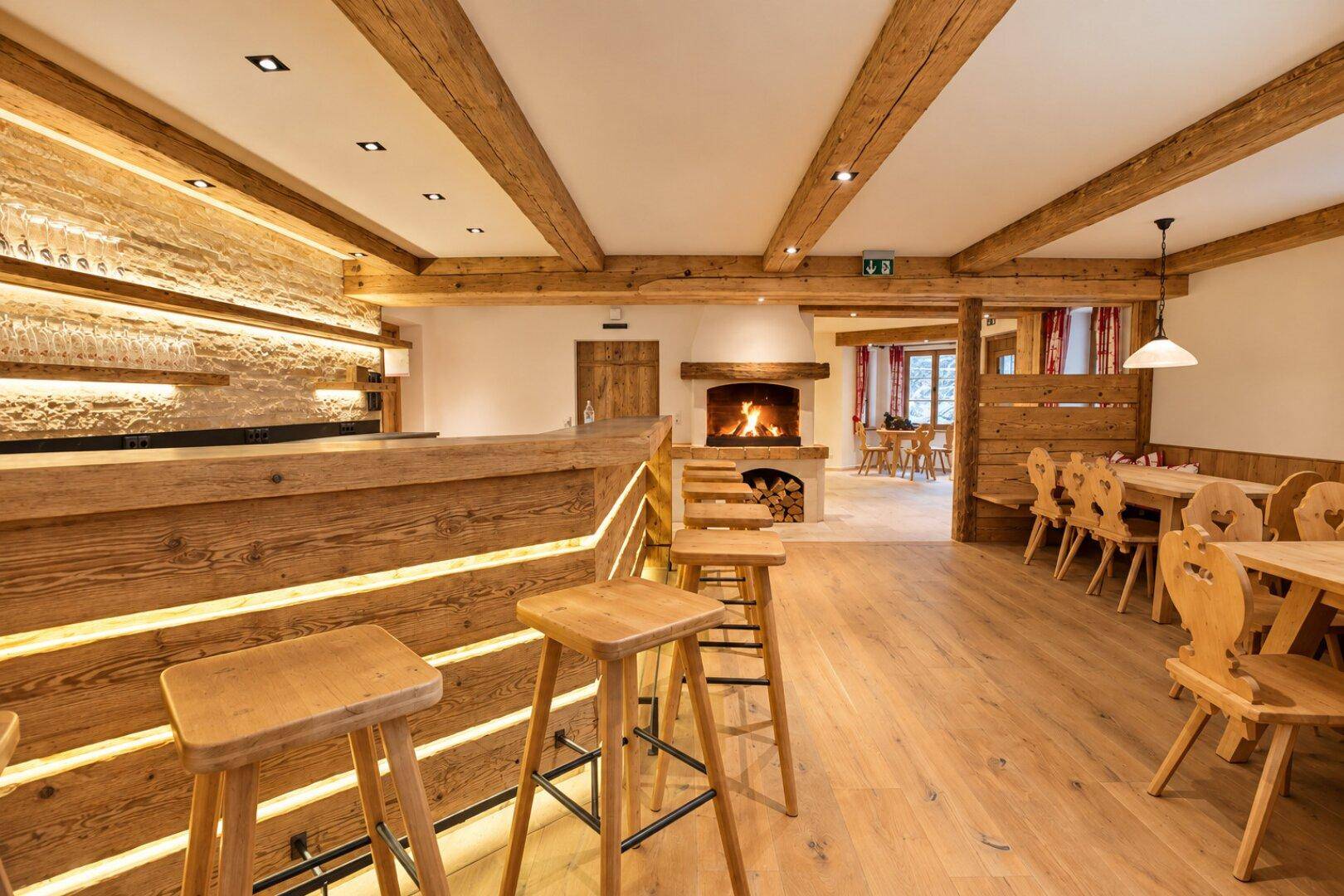 Rustikale Bar mit Holzhockern und Blick auf einen gemütlichen Kaminbereich im Hintergrund.