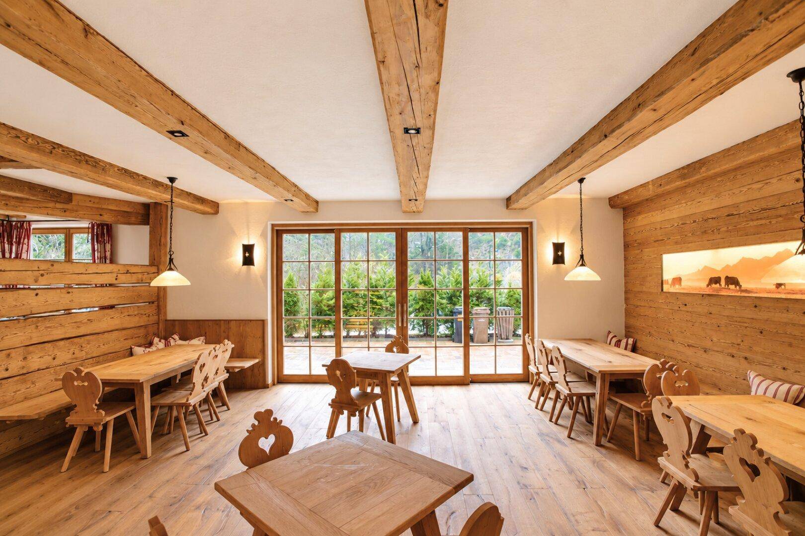 Gemütlicher Speisesaal mit Holztischen, Bänken und sichtbaren Deckenbalken, hell durch große Fenster.