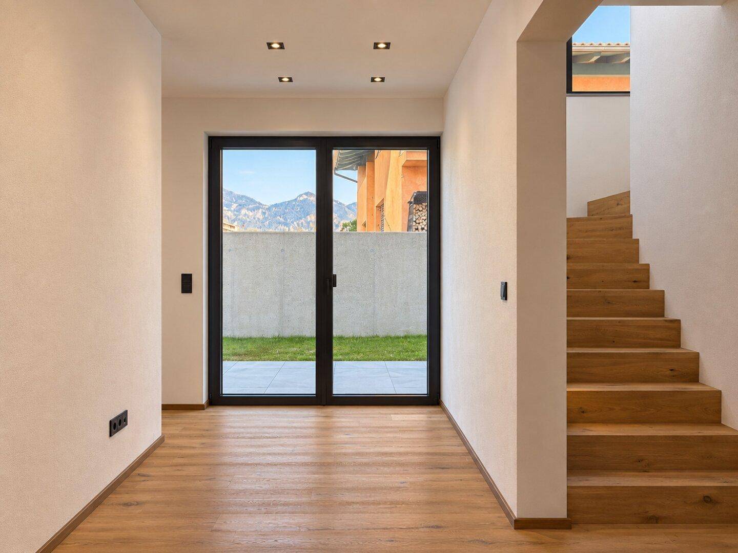 Eingangsbereich mit Holztreppe und großer Glastür, die Zugang zum Garten und Bergblick bietet.