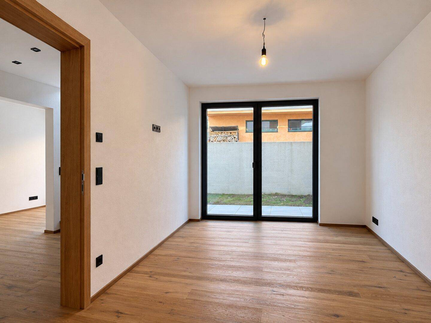 Leeres Zimmer mit Holzboden und großer Fensterfront, die einen Blick auf den Außenbereich ermöglicht.