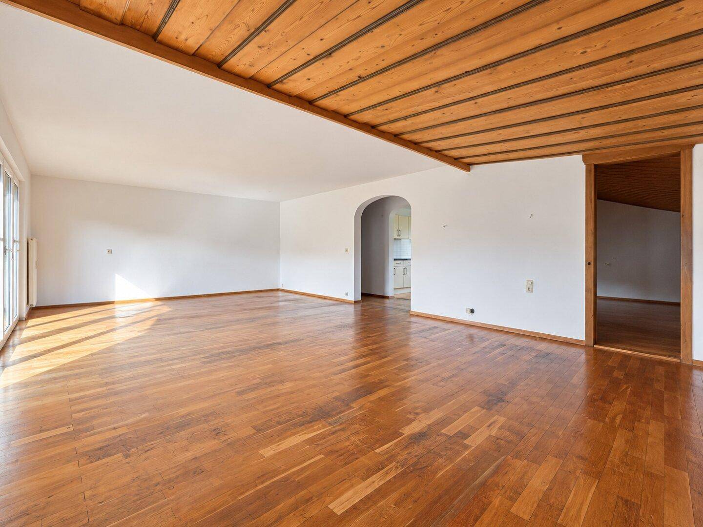 Geräumiges Wohnzimmer mit Holzboden, Holzbalkendecke und Zugang zu einem weiteren Raum.