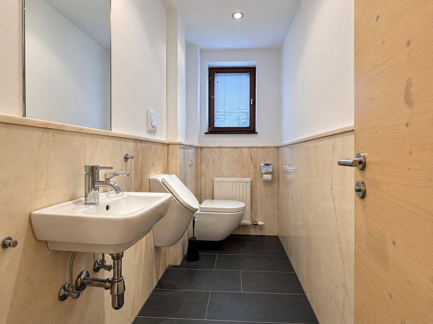 Modernes Gäste-WC mit Wand-WC, Waschbecken und Fenster, halbhoch gefliest.