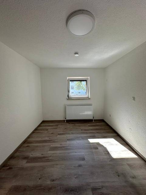 Leerer Raum mit hellem Laminatboden und einem Fenster, das Tageslicht hereinlässt.