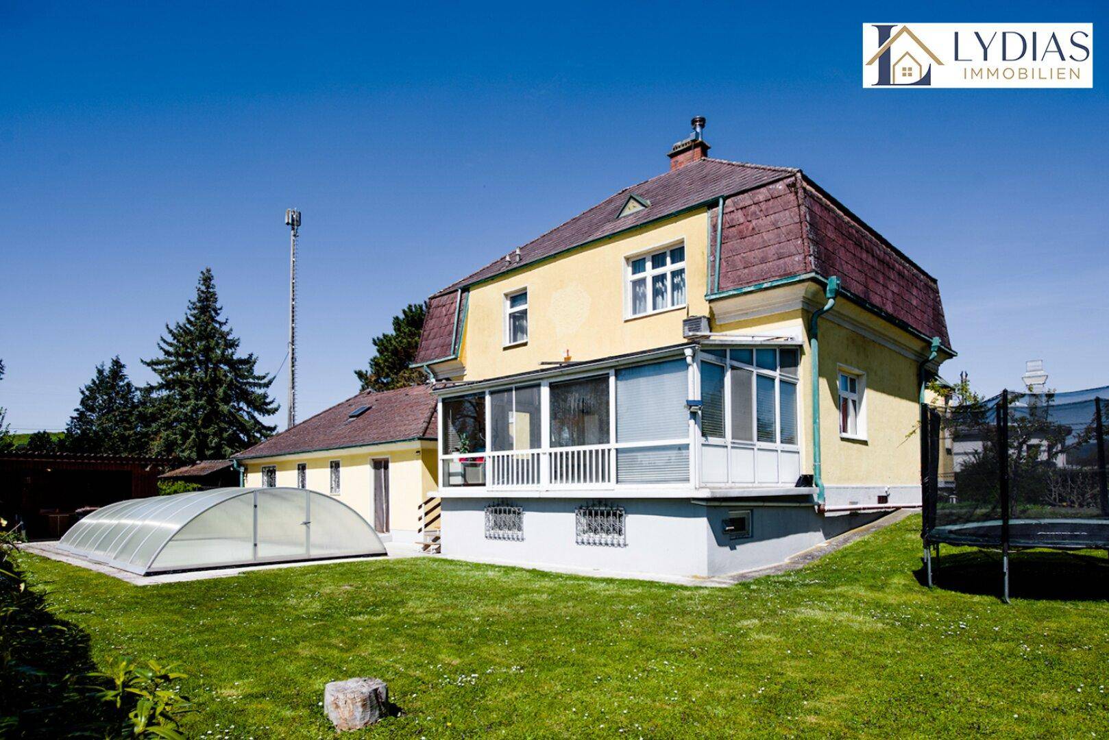 Großer Gartenbereich mit Rasenfläche, Trampolin und überdachtem Pool auf sonnigem Grundstück.