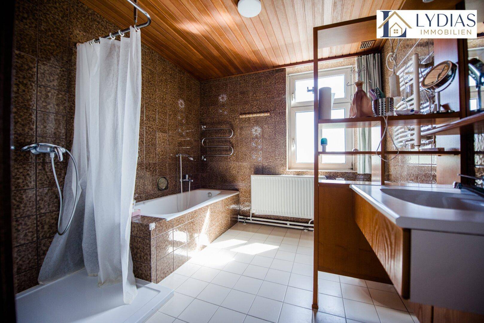Badezimmer mit braunen Fliesen, Badewanne, Dusche und Fenster für natürliche Belüftung.