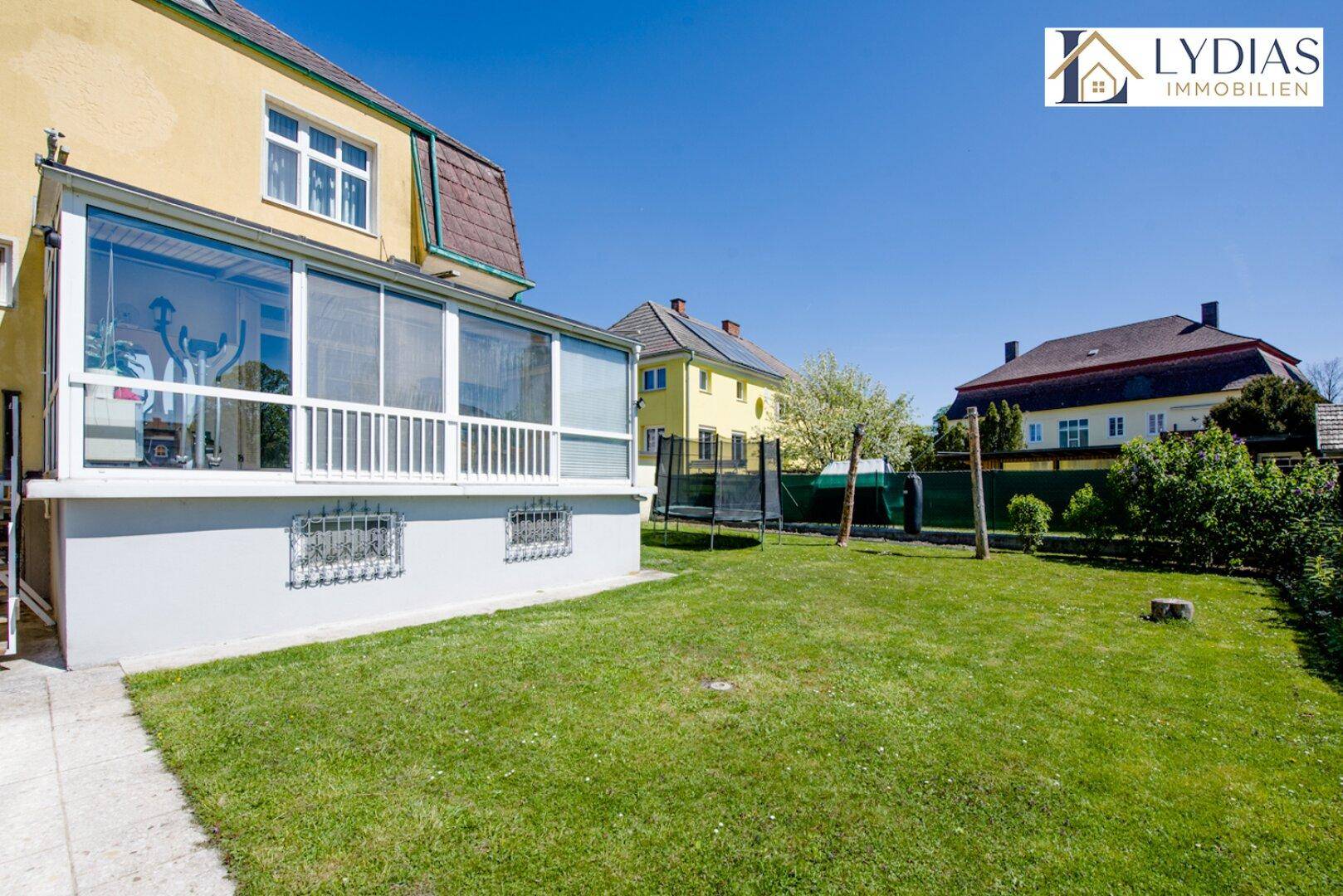 Großer Garten mit Rasenfläche, Trampolin und einem verglasten Wintergarten am Haus.