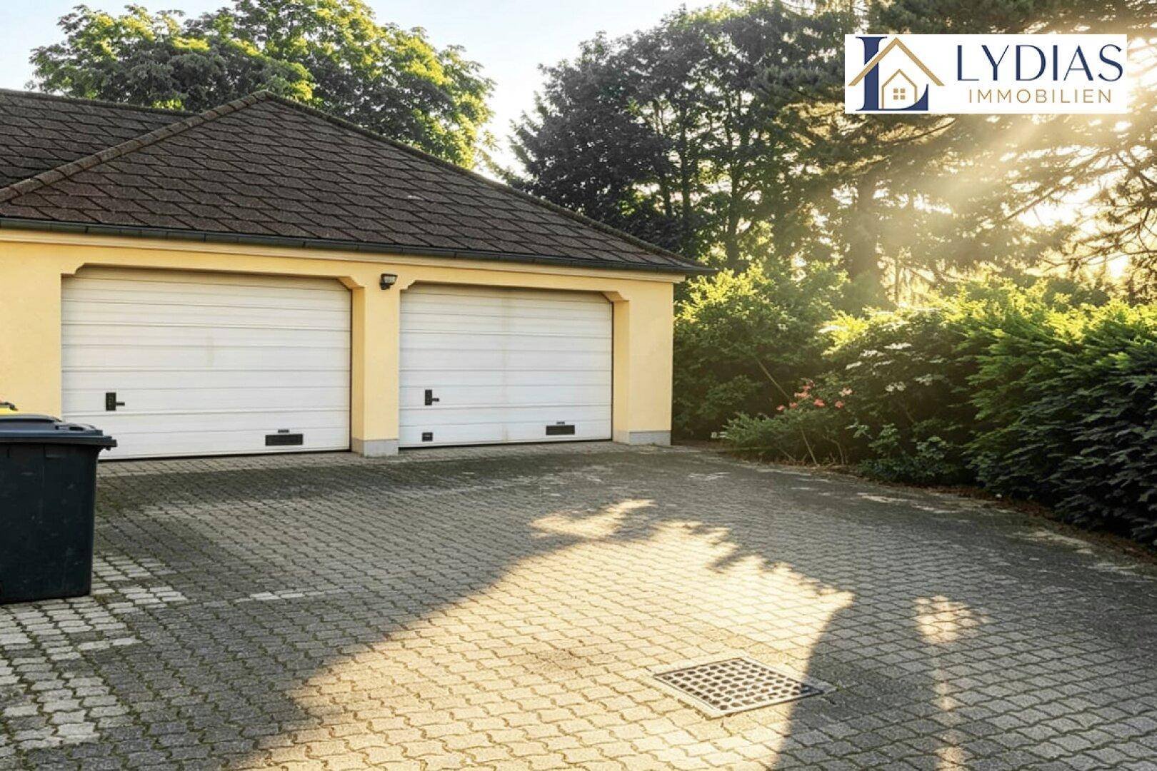 Doppelgarage mit zwei weißen Toren und gepflasterter Einfahrt, umgeben von Bäumen und Sträuchern.