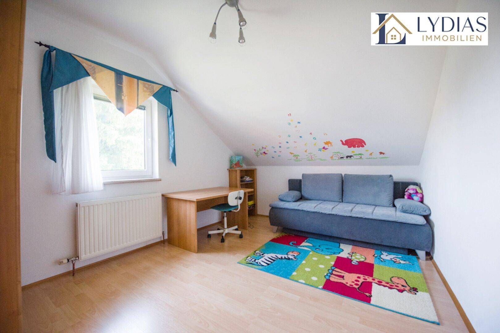 Helles Zimmer im Dachgeschoss mit Schreibtisch, Sofa und farbenfrohem Teppich.