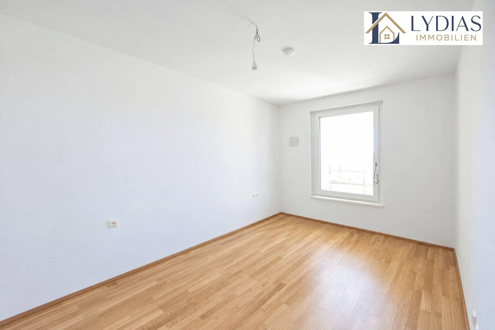 Leeres Schlafzimmer mit hellem Holzboden und einem großen Fenster für viel Tageslicht.