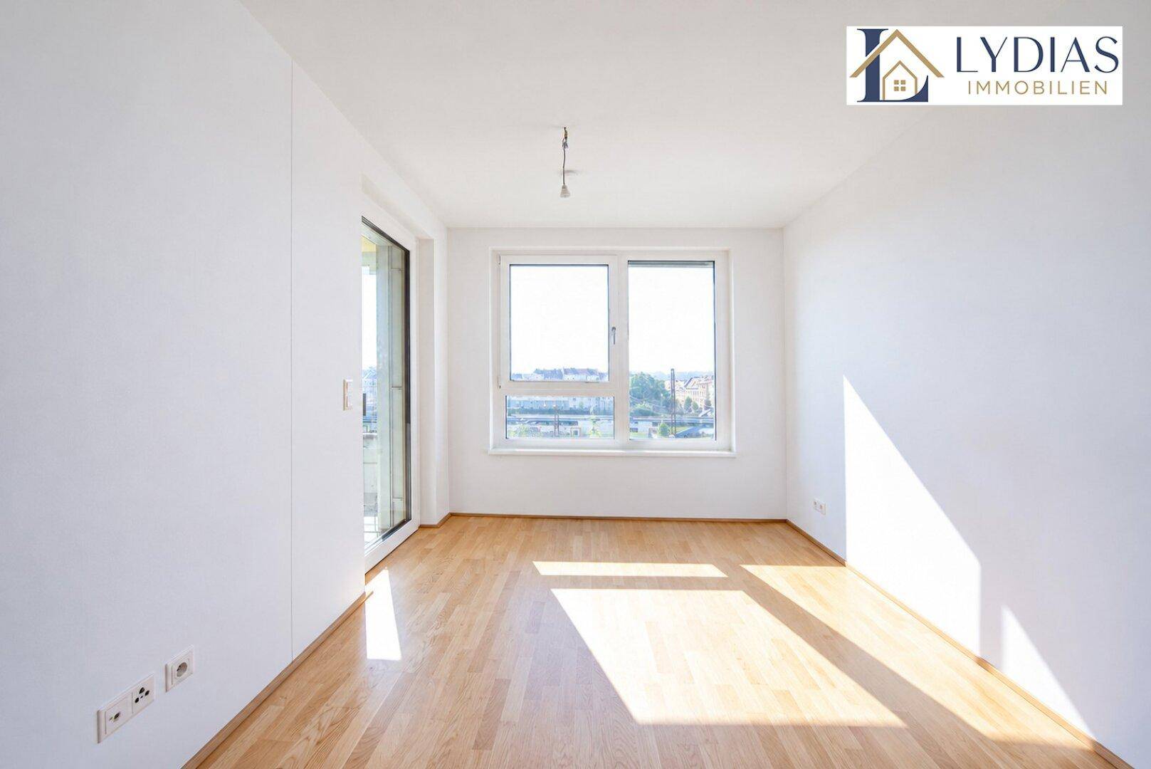 Leeres Wohnzimmer mit hellem Holzboden, großen Fenstern und viel Tageslicht.